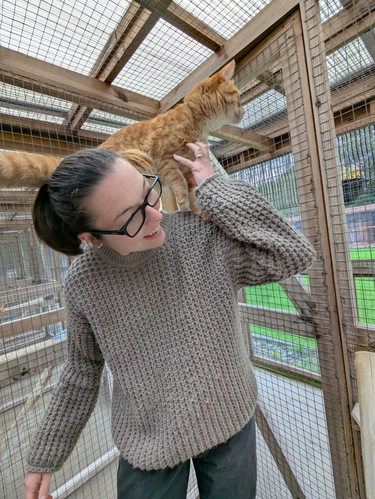 A woman with short dark hair, glasses, and a beige sweater holding a ginger cat on her shoulder inside a wooden and wire enclosure.