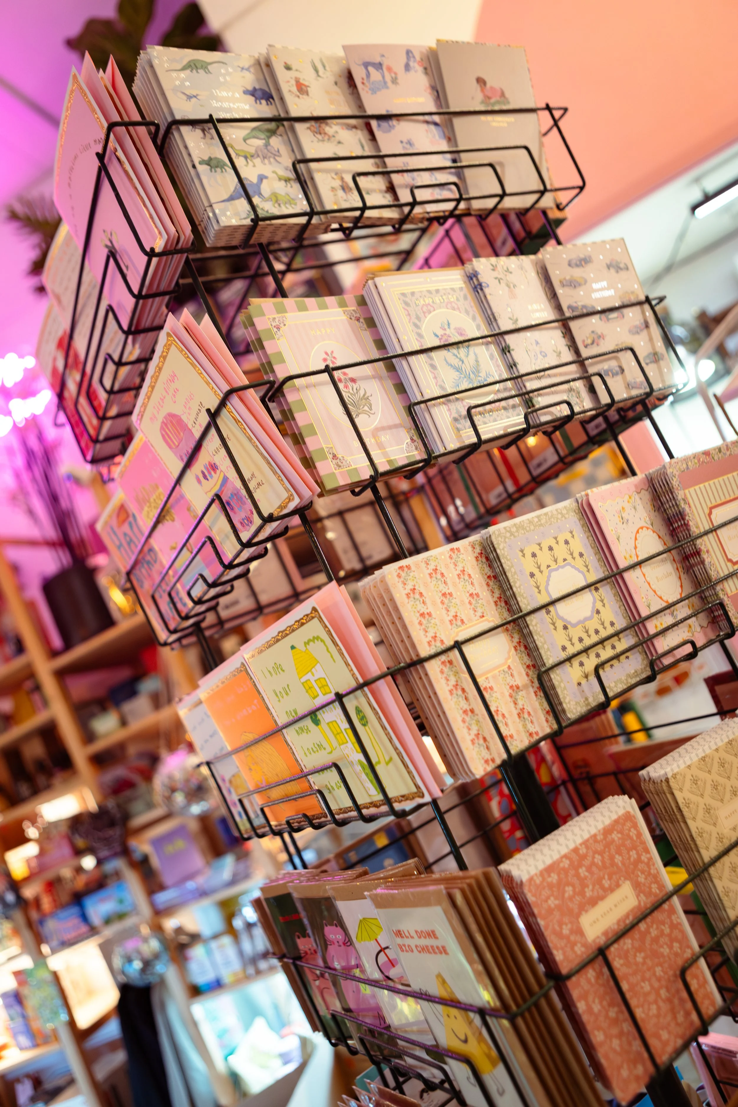 A black metal rack displaying various colorful greeting cards inside a store with a pink and purple wall background.