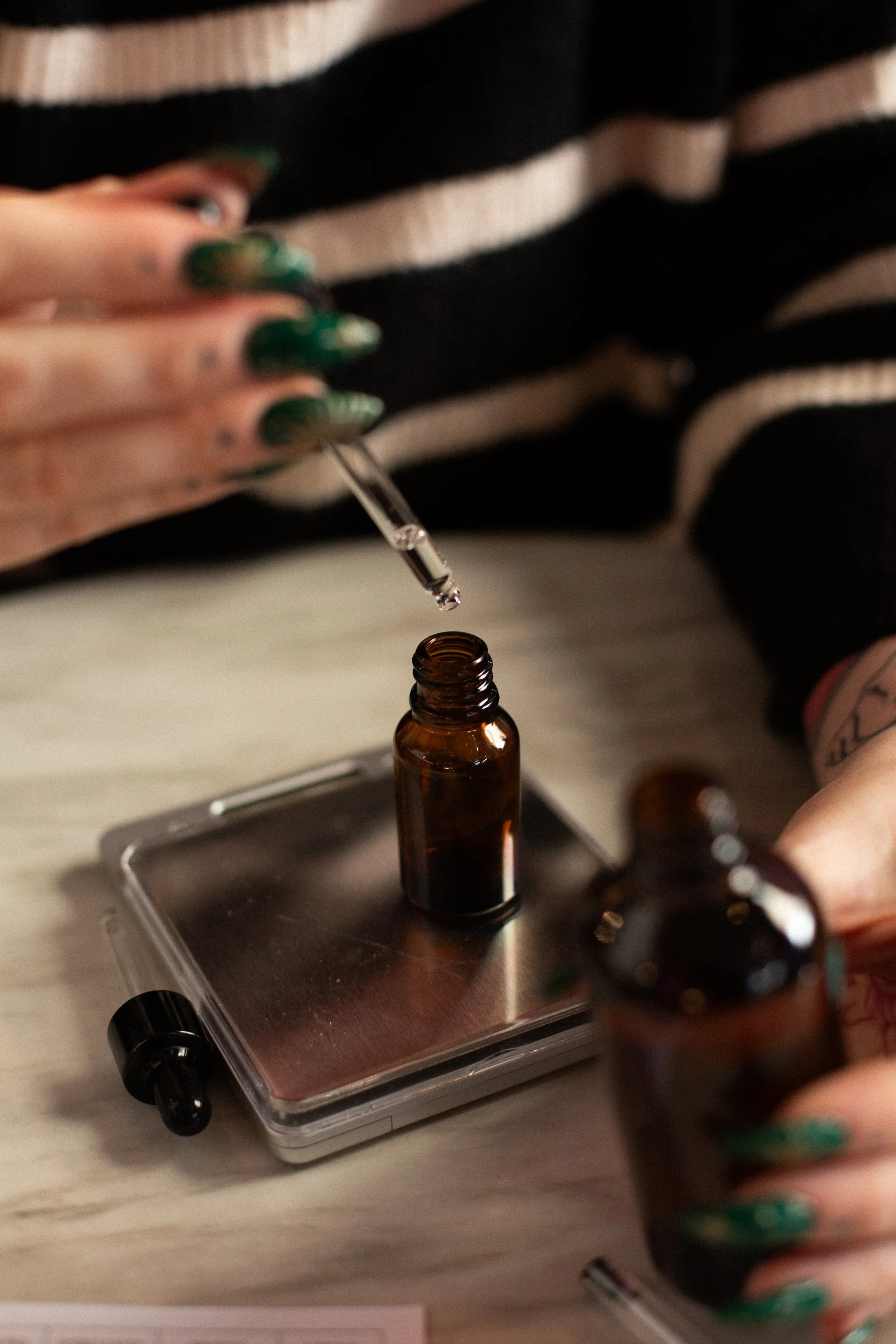 Person with tattooed hands and green nails using a dropper to transfer a liquid into a small brown glass bottle on a digital scale.