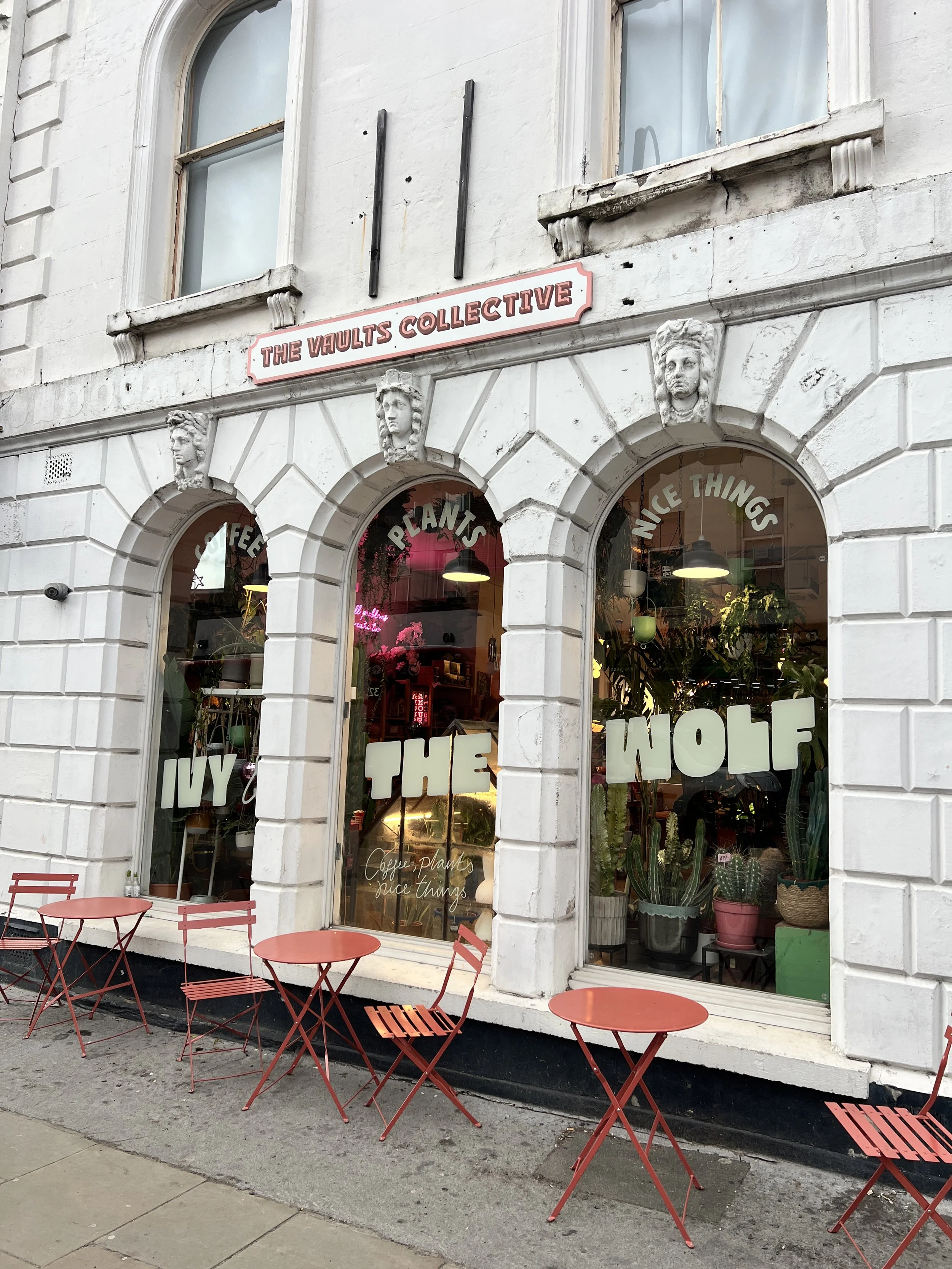 The storefront of The Vaults Collective, a shop with large arched windows displaying plants and signs, with red outdoor tables and chairs outside.