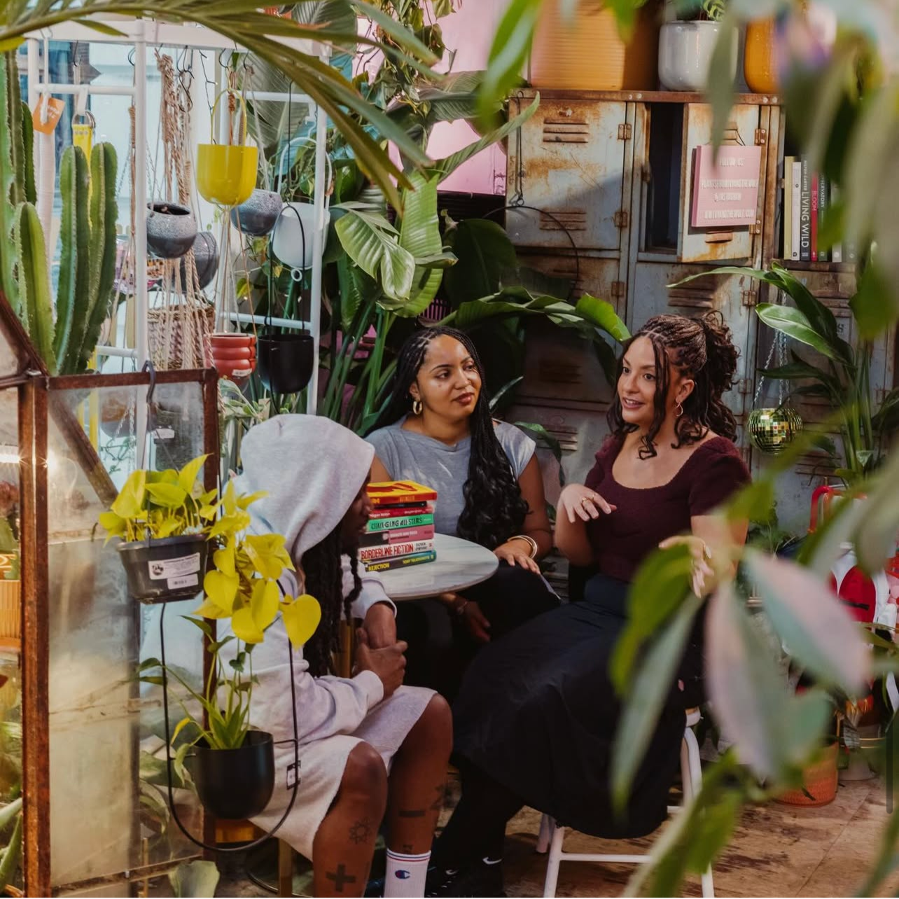 Four women sitting around a small table in a lush, decorated indoor space with plants, lockers, and hanging pots, engaged in conversation.