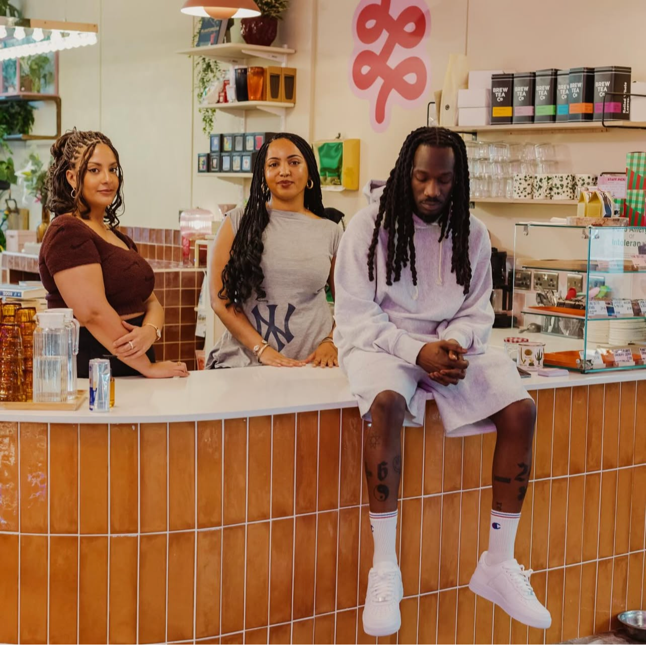 Three people at a cafe counter, two women standing and a man sitting on the counter. The women are smiling, while the man looks down with his hands clasped.
