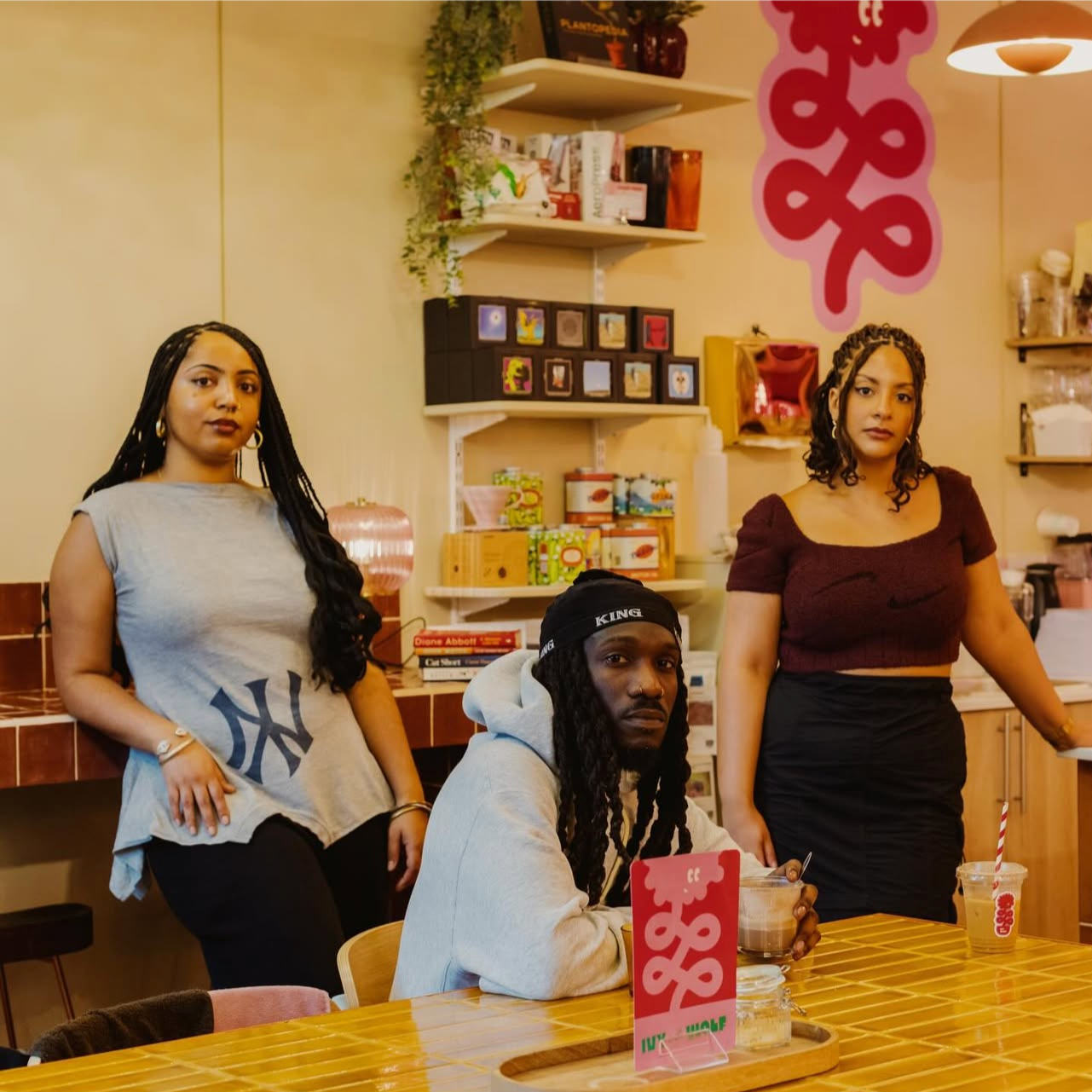 Three young adults sitting and standing in a cafe, with shelves and colorful decor in the background.