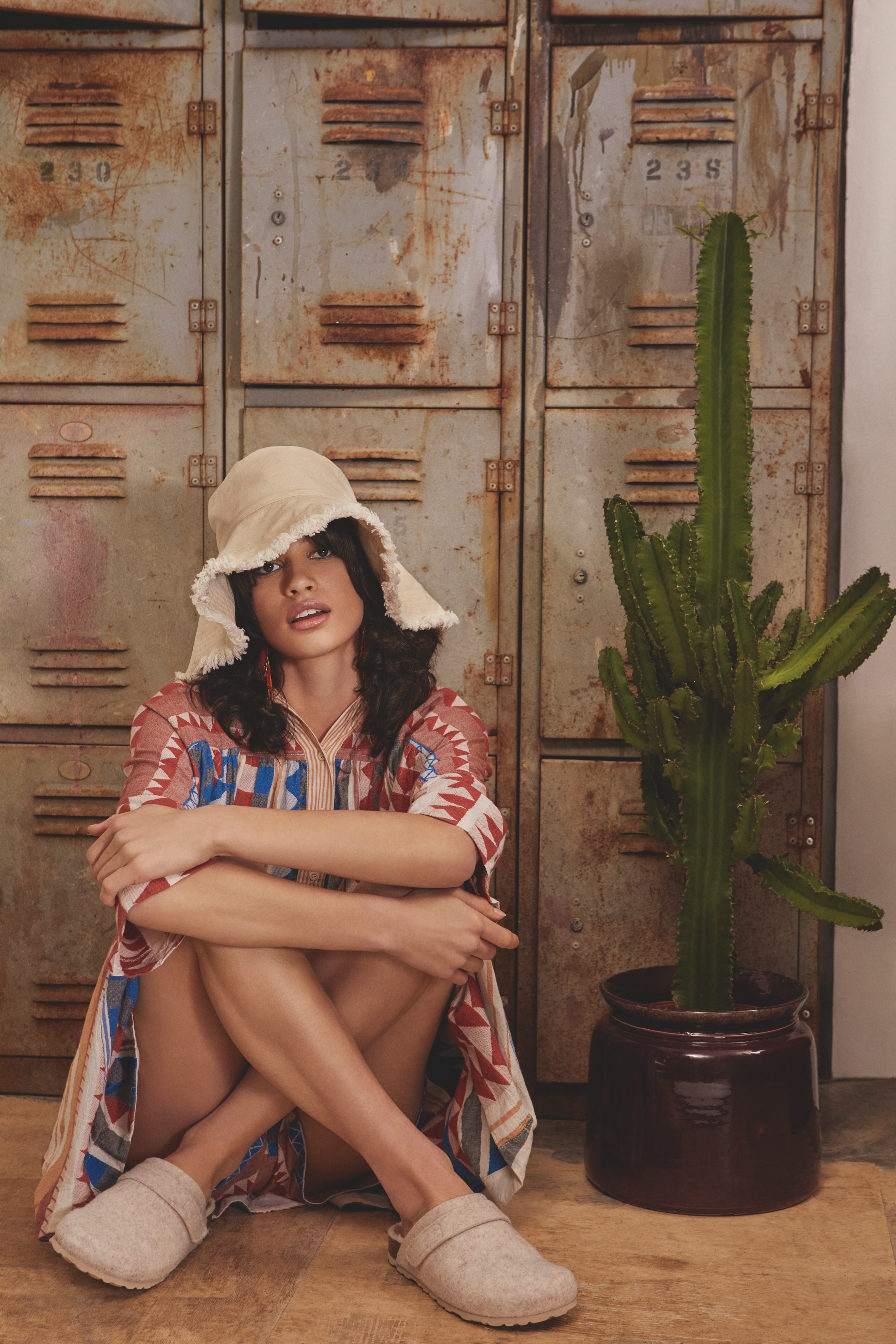 A woman sitting on the floor next to a large potted cactus with rusty lockers in the background.