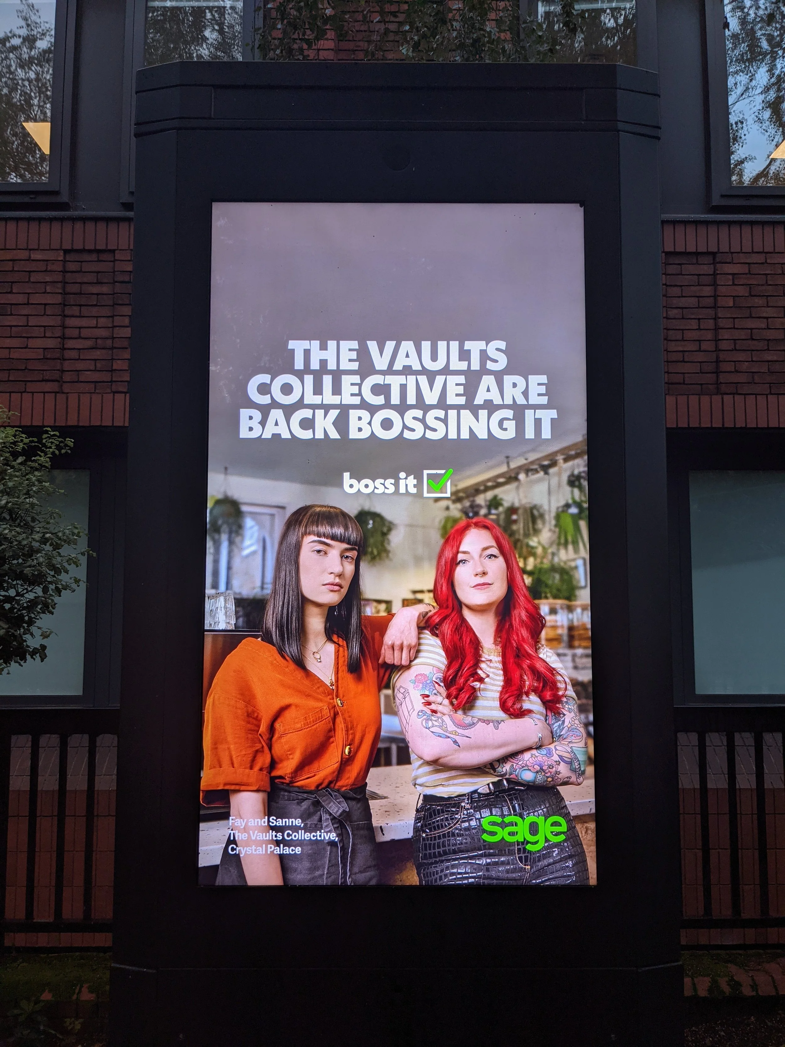 Poster featuring two women with the text 'The vaults collective are back bossing it' and the logo of Sage at the bottom right. One woman has straight dark hair and an orange top, the other has wavy red hair and tattoos on her arms, standing in an ind