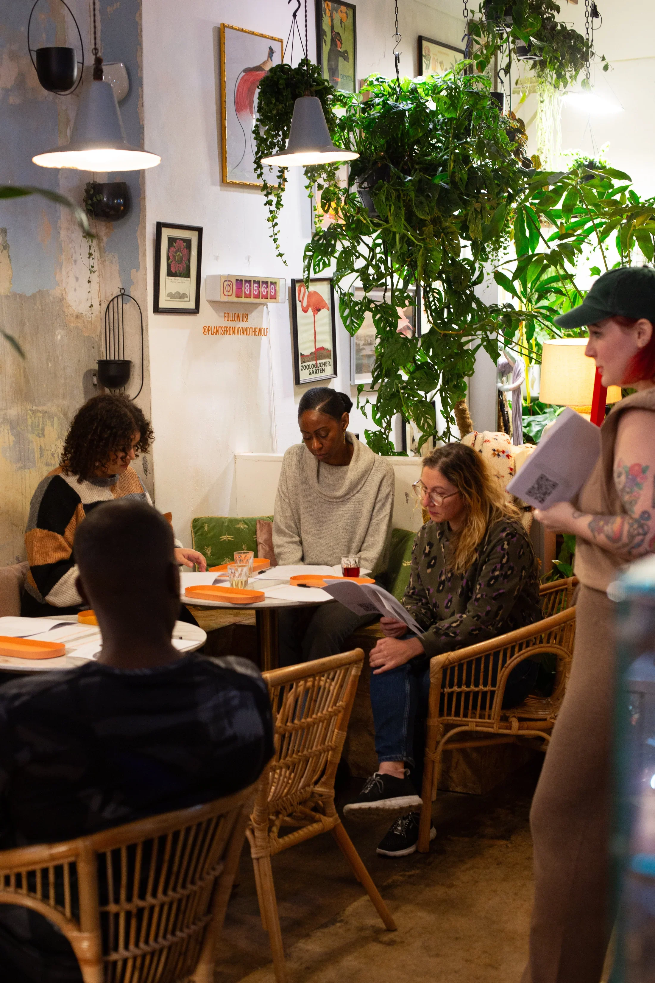 A group of five people sitting around a table in a cozy, decorated cafe, with plants, framed pictures, and hanging lights on the wall.