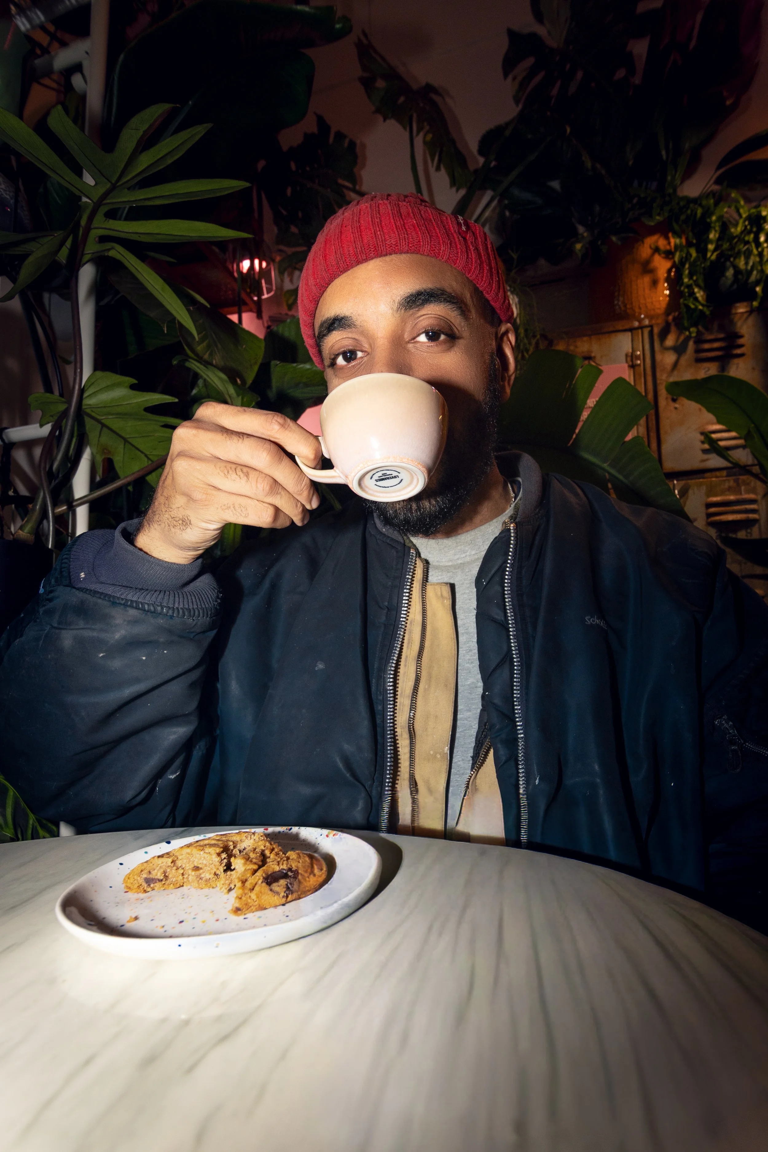 Man wearing a red beanie and black jacket drinking from a white mug, seated at a table with a plate of cookies, surrounded by large green plants and cozy lighting.