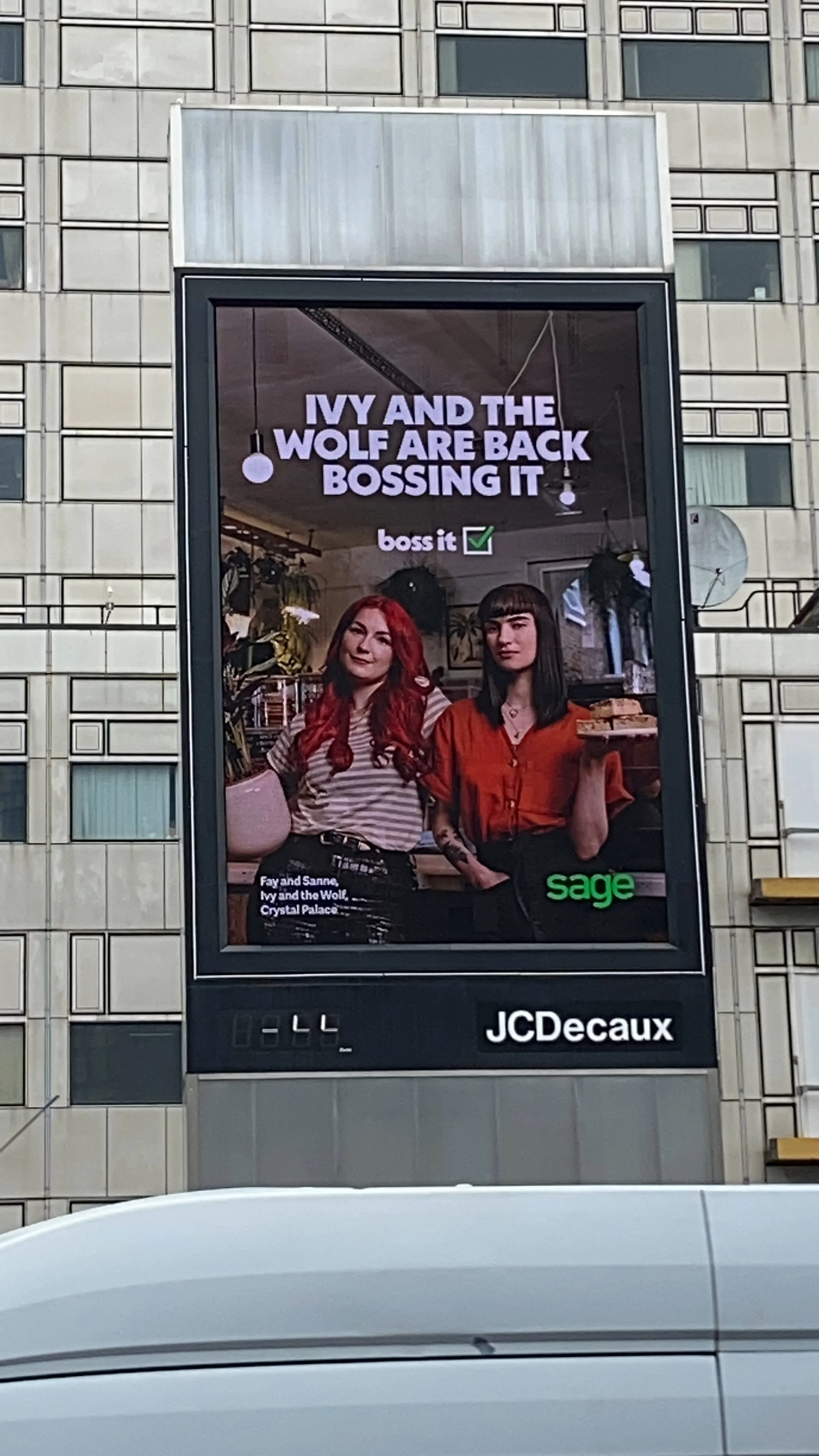 Billboard advertisement featuring two women in a café; one with red hair wearing a striped t-shirt, the other with black hair in an orange blouse holding a tray; text overlayed reads, 'IVY AND THE WOLF ARE BACK BOSSING IT' and 'sage' appears at the b
