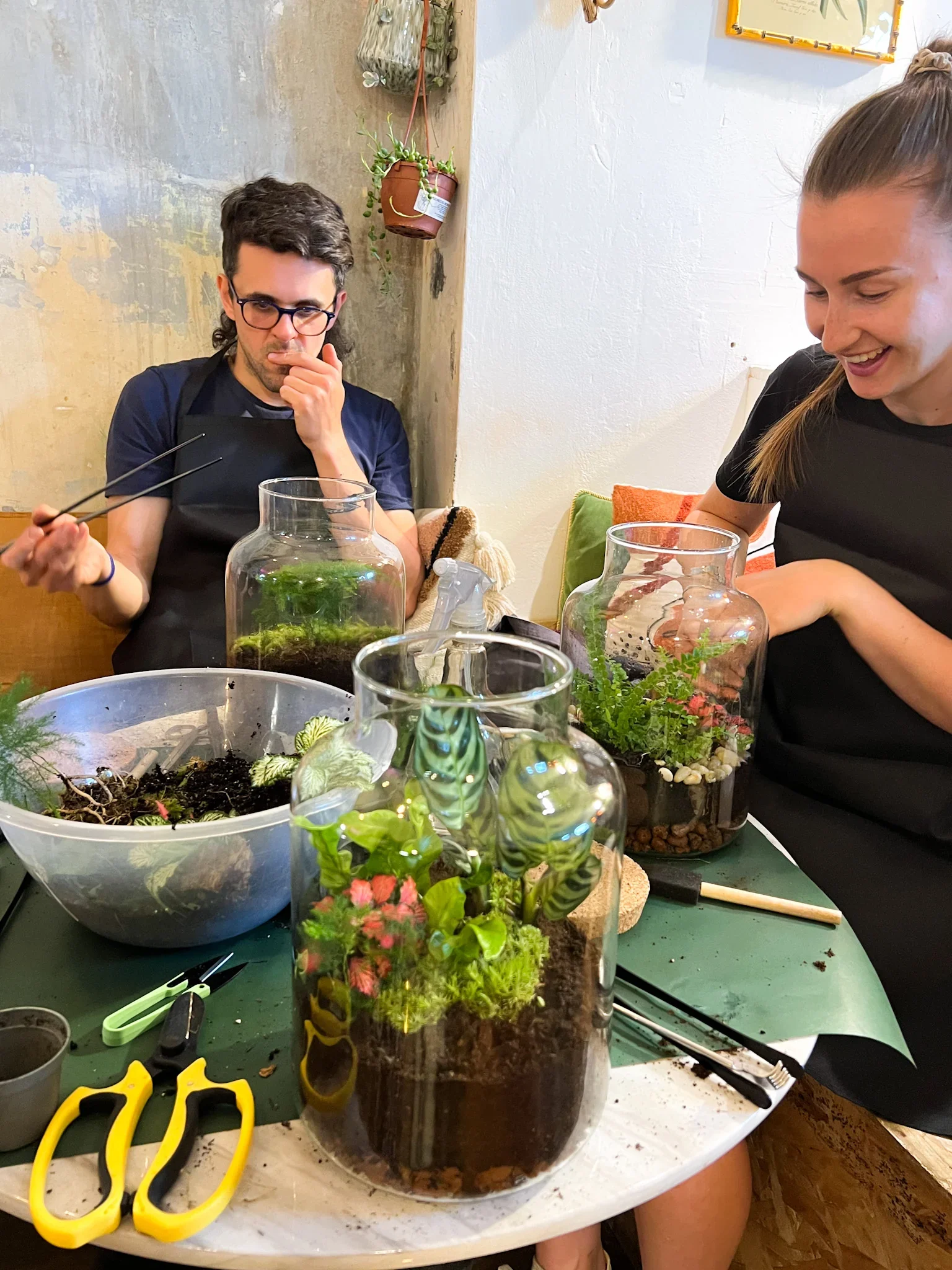 Two people working on glass terrariums with plants on a table, surrounded by gardening tools.