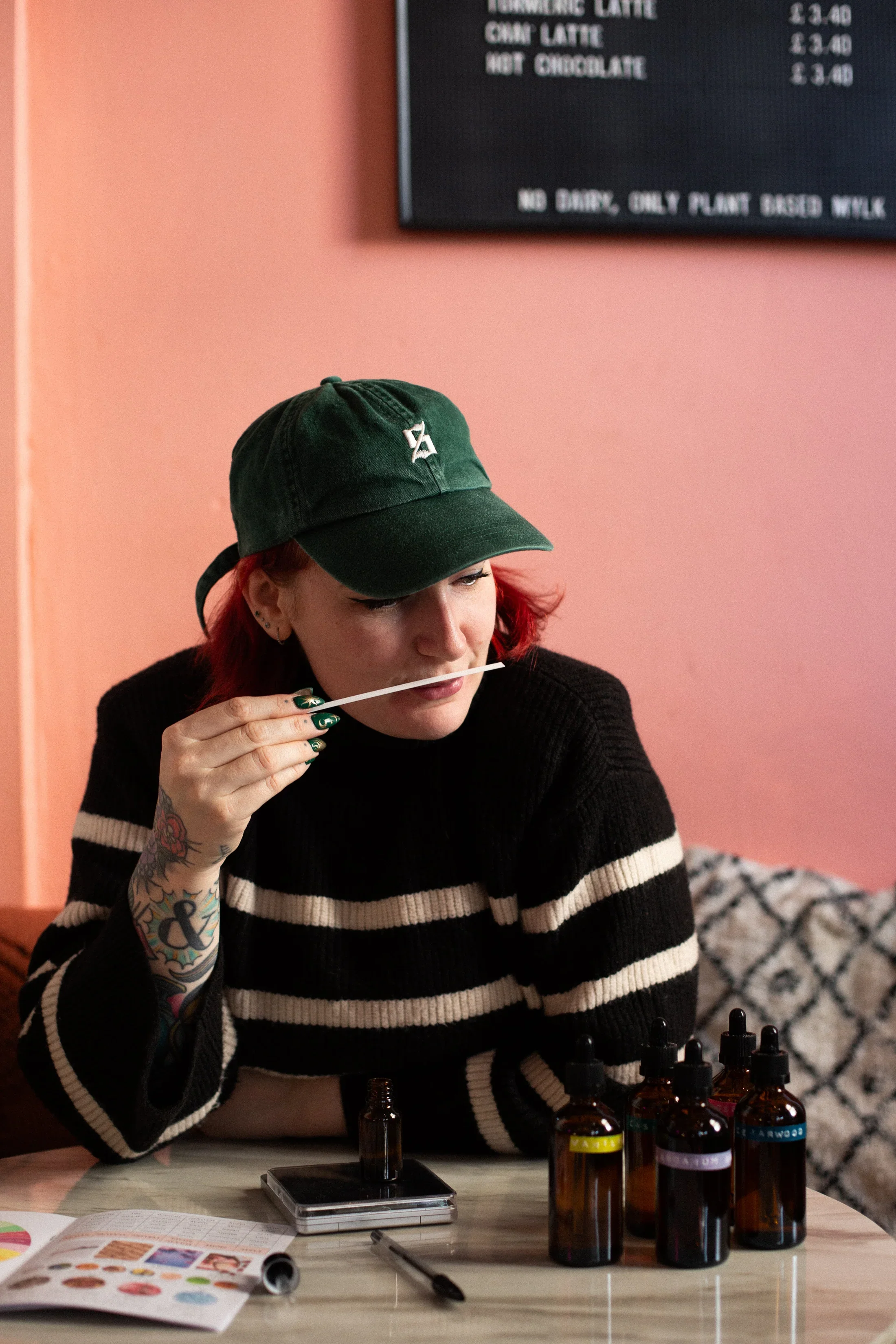 Woman with red hair and tattoos, wearing a green cap and black-and-white striped sweater, holds a dropper near her lips while sitting at a table with essential oil bottles, a pen, a phone, and a pamphlet, inside a room with pink walls and a patterned cushion.