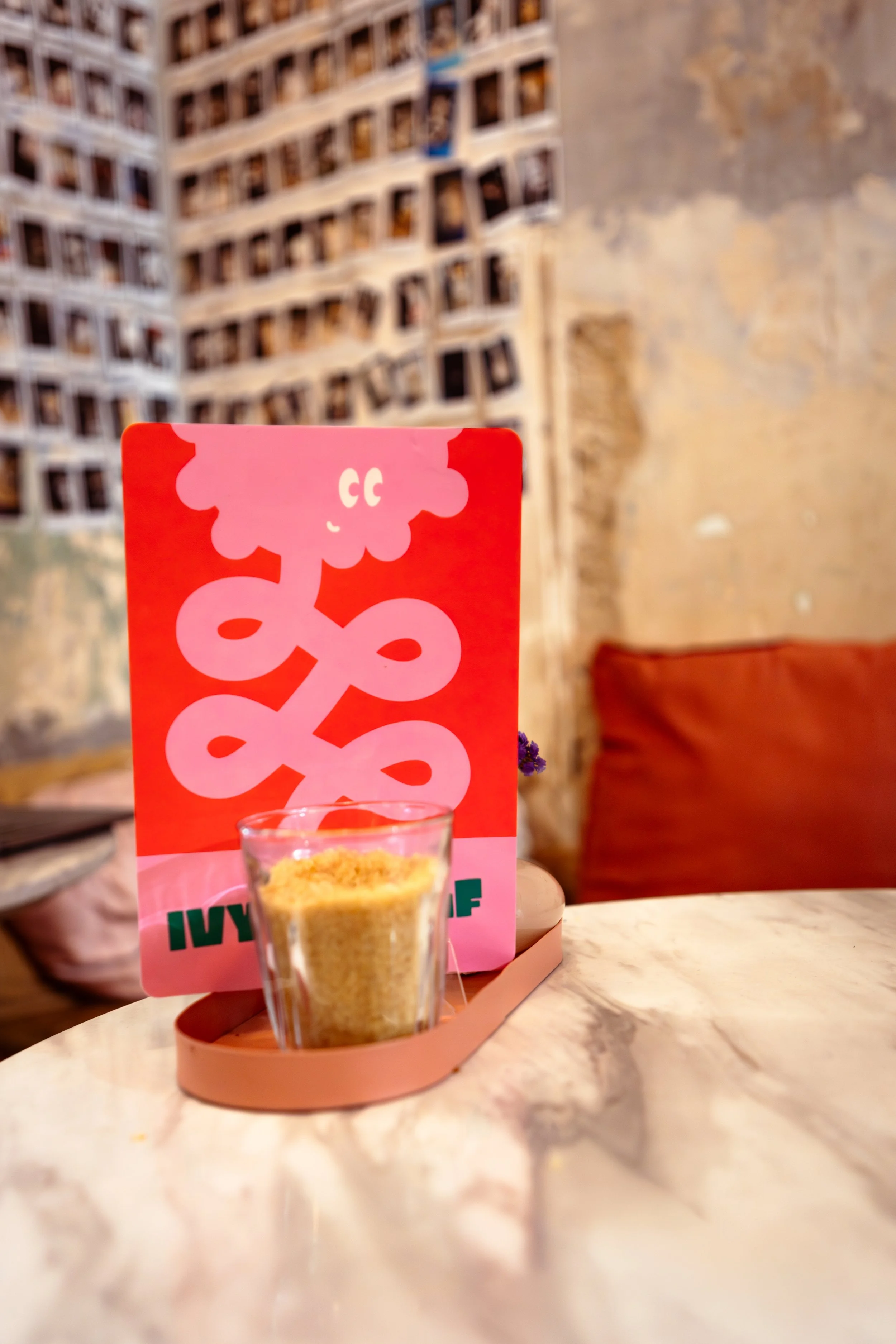 A glass of yellow sand on a beige table in front of a pink folder with a cartoon pink cloud and a white abstract pattern. The folder has English and Spanish text "Ivy City". The background features a beige wall with peeling paint and a red cushion.