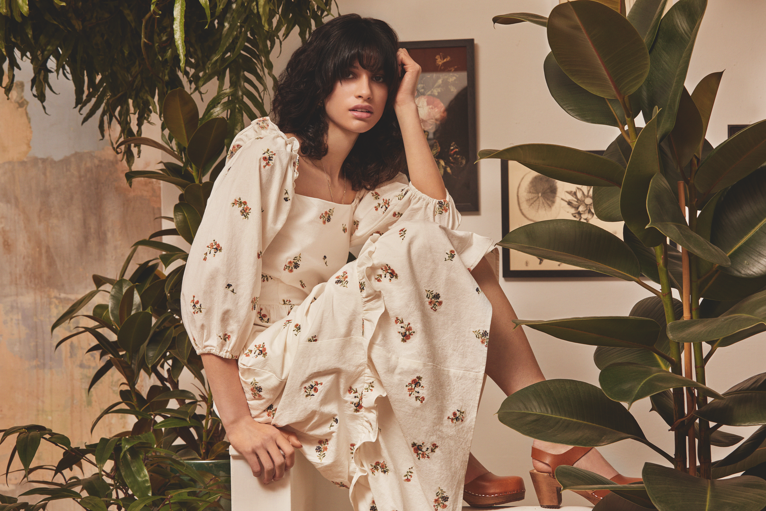 A woman with dark curly hair wearing a cream-colored floral dress, sitting among large green plants and framed botanical artwork in a cozy interior.