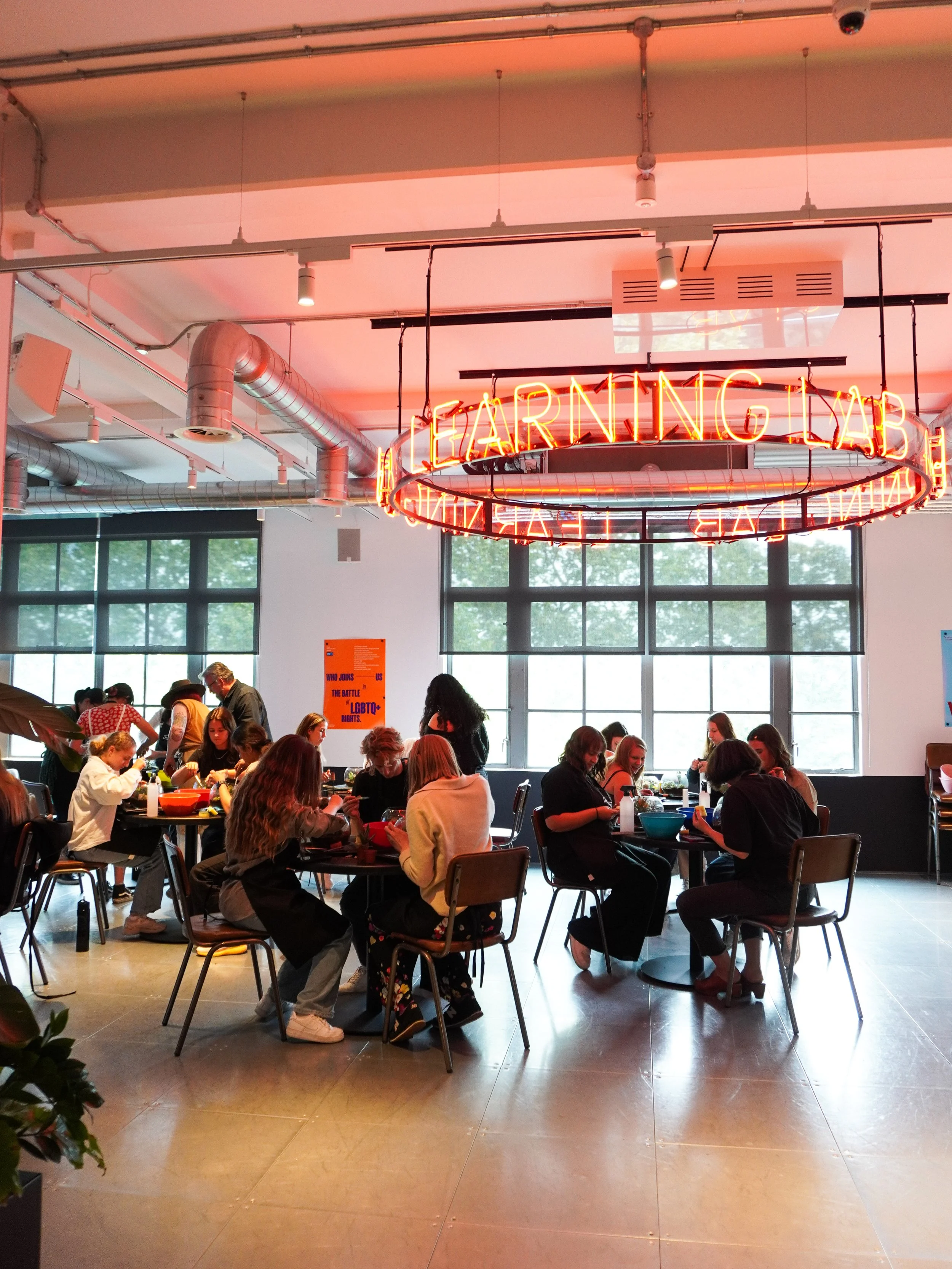 People sitting at tables in a modern, brightly lit learning club with large windows, exposed ductwork, and a neon circular sign reading 'Learning Club'.