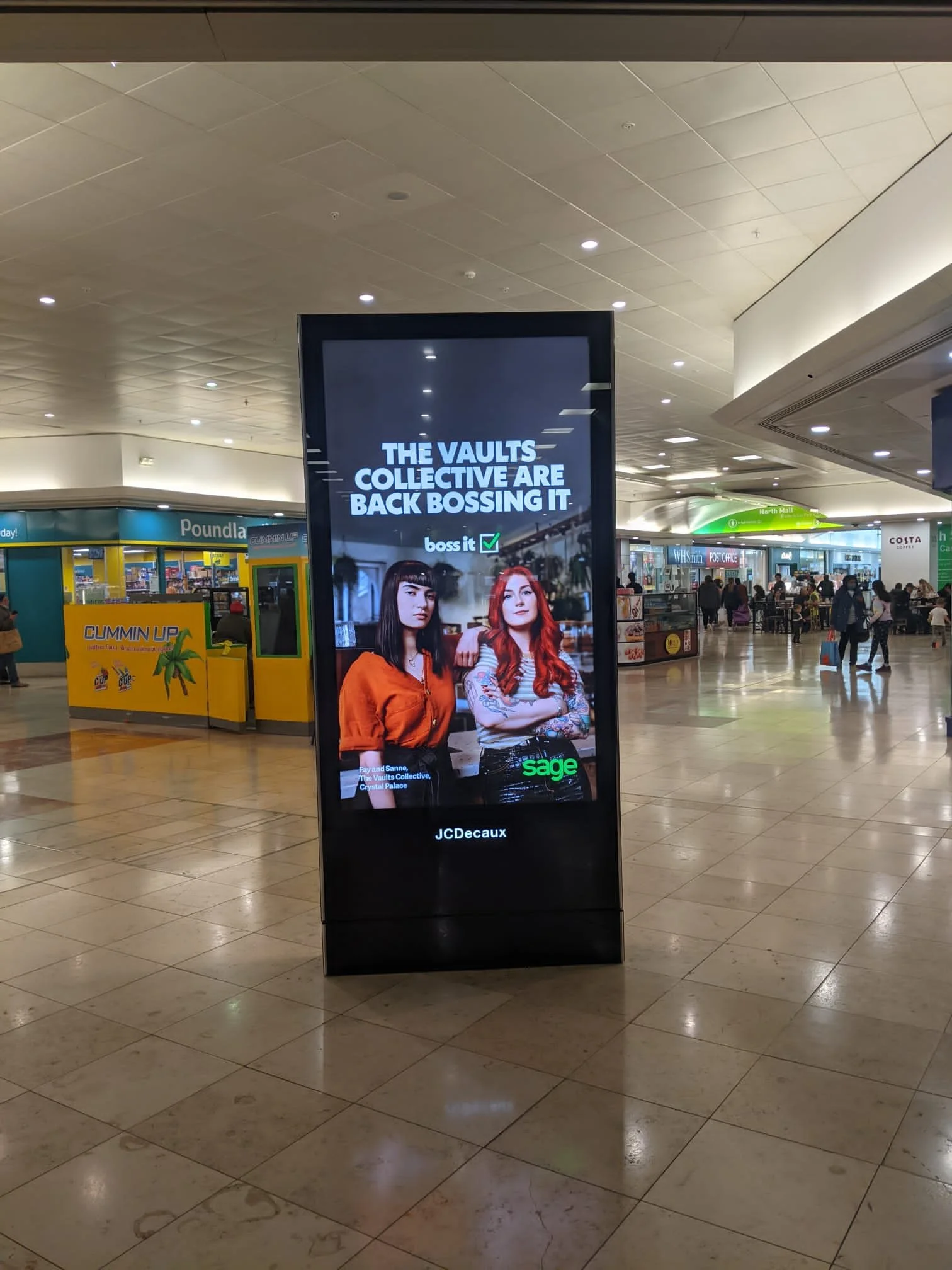 Digital advertisement in a shopping mall featuring two women with the text 'The vaults collective are back bossing it' and logos of Sage and JCDecaux.