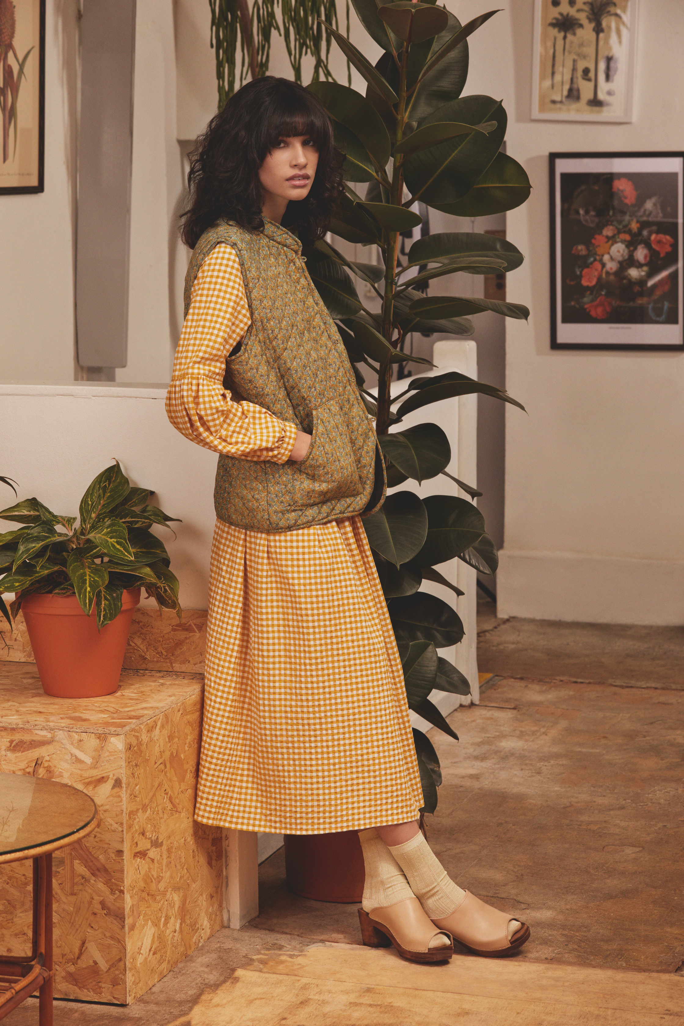 Woman standing in a cozy indoor space with plants and framed artwork, wearing a yellow and checkered skirt, patterned vest, and beige open toe shoes.