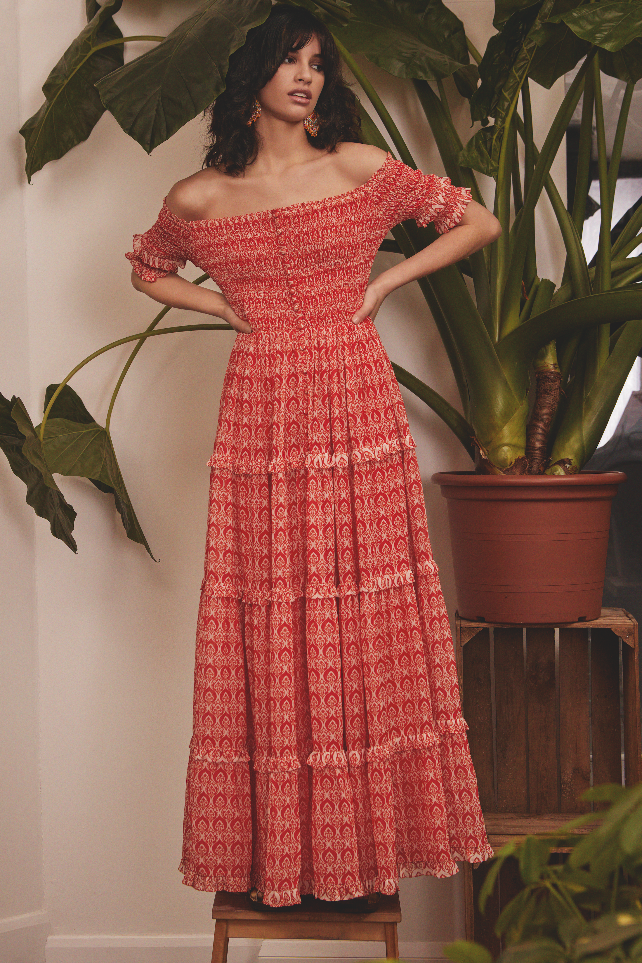 Woman wearing a red patterned off-shoulder maxi dress with ruffles, standing in front of a plant and a white wall.