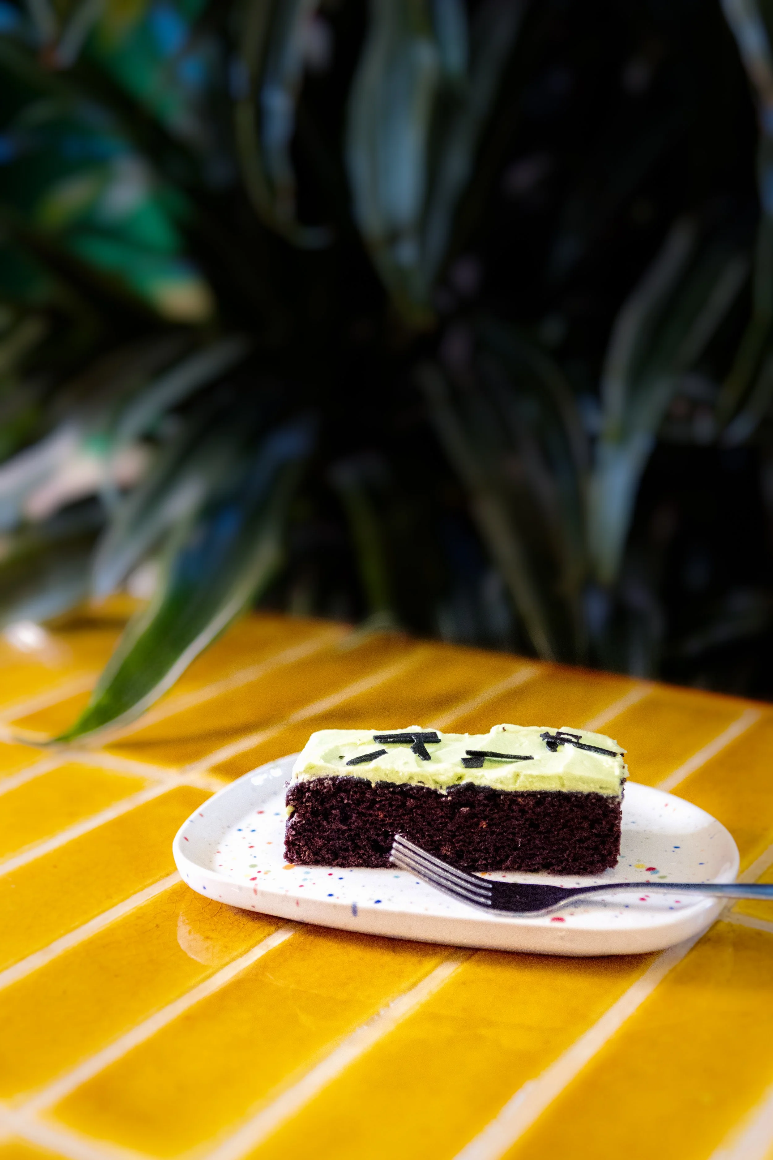 A slice of chocolate cake with green frosting and chocolate sprinkles on top, served on a white speckled plate on a yellow-tiled table with green plants in the background.