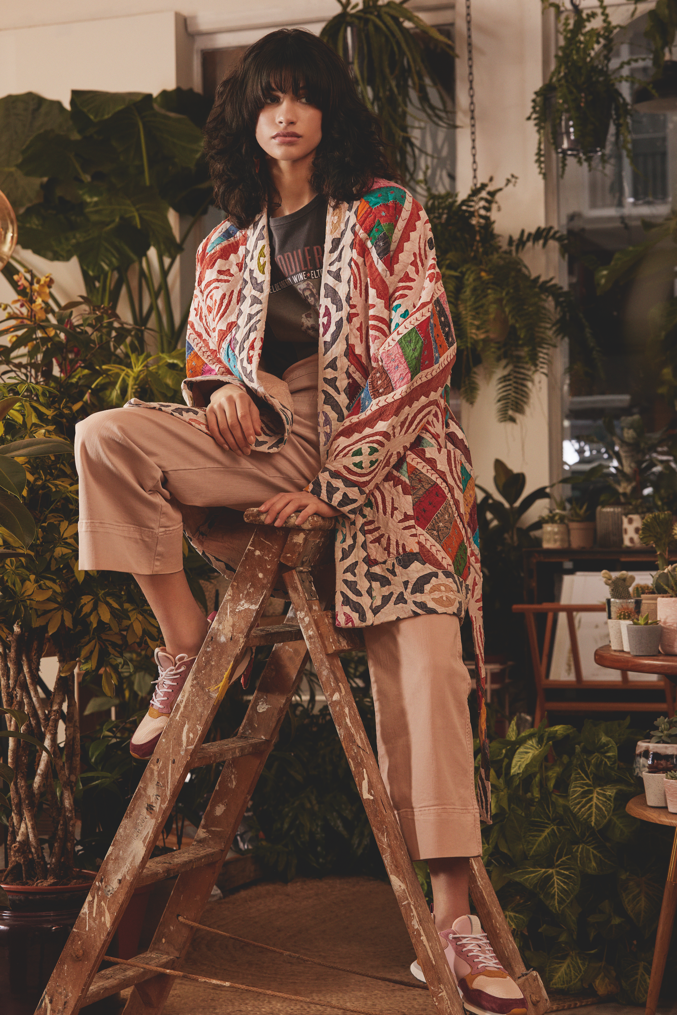 A woman with dark, curly hair sits on a wooden ladder in a room filled with green plants, wearing beige pants, pink sneakers, and a colorful, patterned coat.