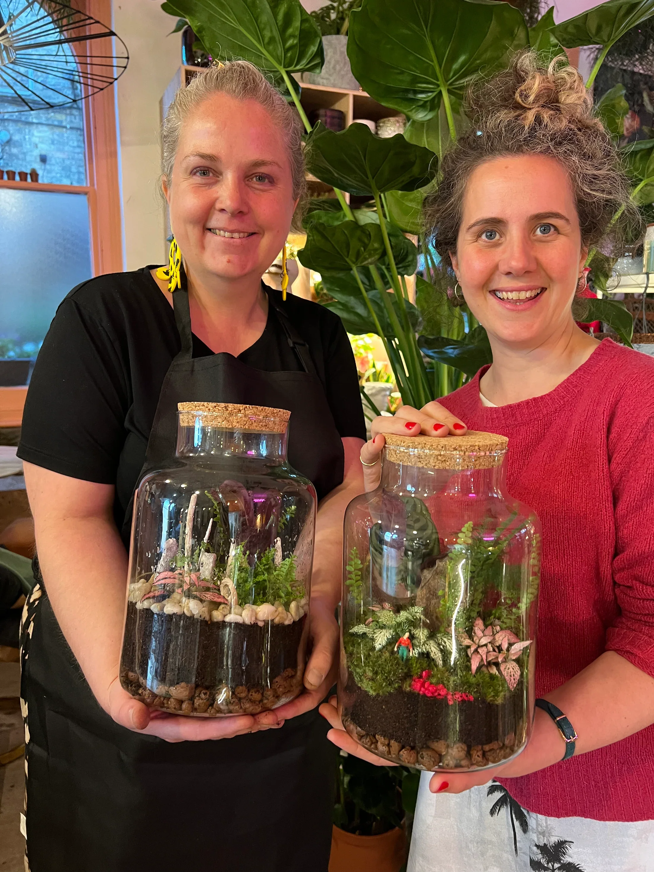 Two women smiling and holding glass terrariums with plants and miniature decorations inside, standing in a room with large green plants and a window in the background.