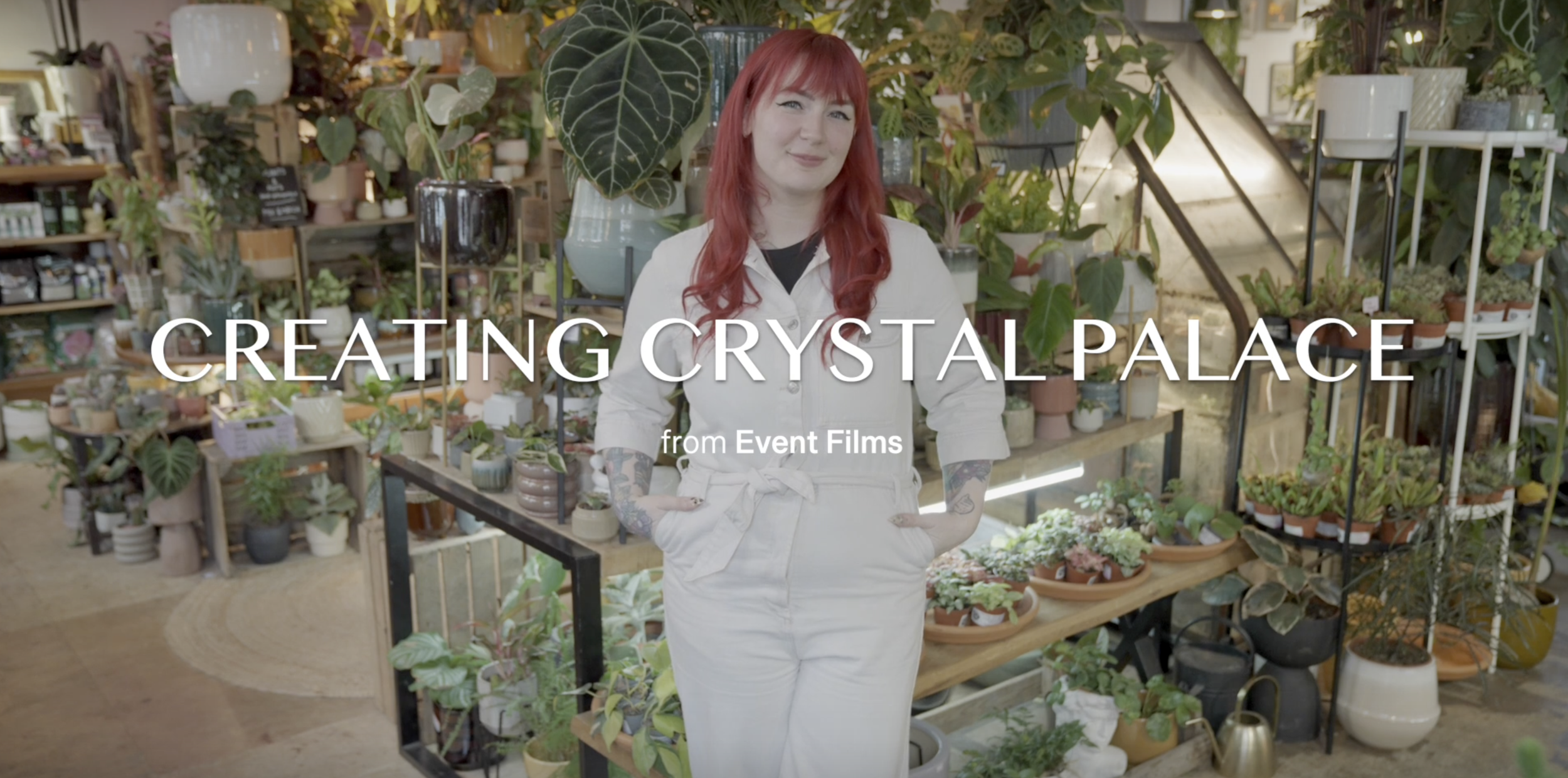 A woman with red hair and tattoos wearing a white jumpsuit, standing in a plant shop surrounded by various potted plants and greenery.