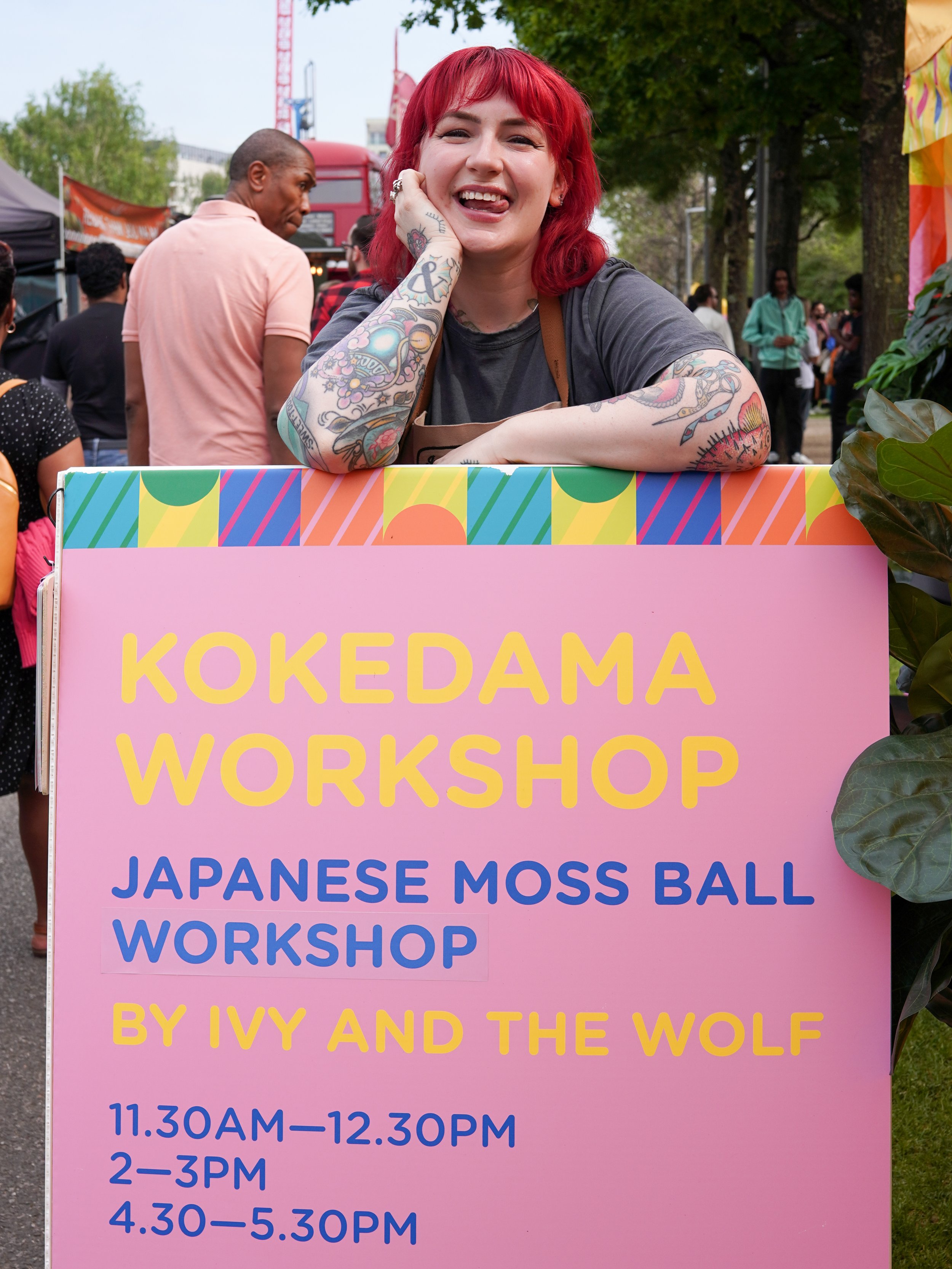 A smiling woman with red hair, tattoos, and a grey t-shirt, leaning on a colorful sign at an outdoor event. The sign advertises a Japanese moss ball workshop by Ivy and the Wolf, taking place at various times during the day.