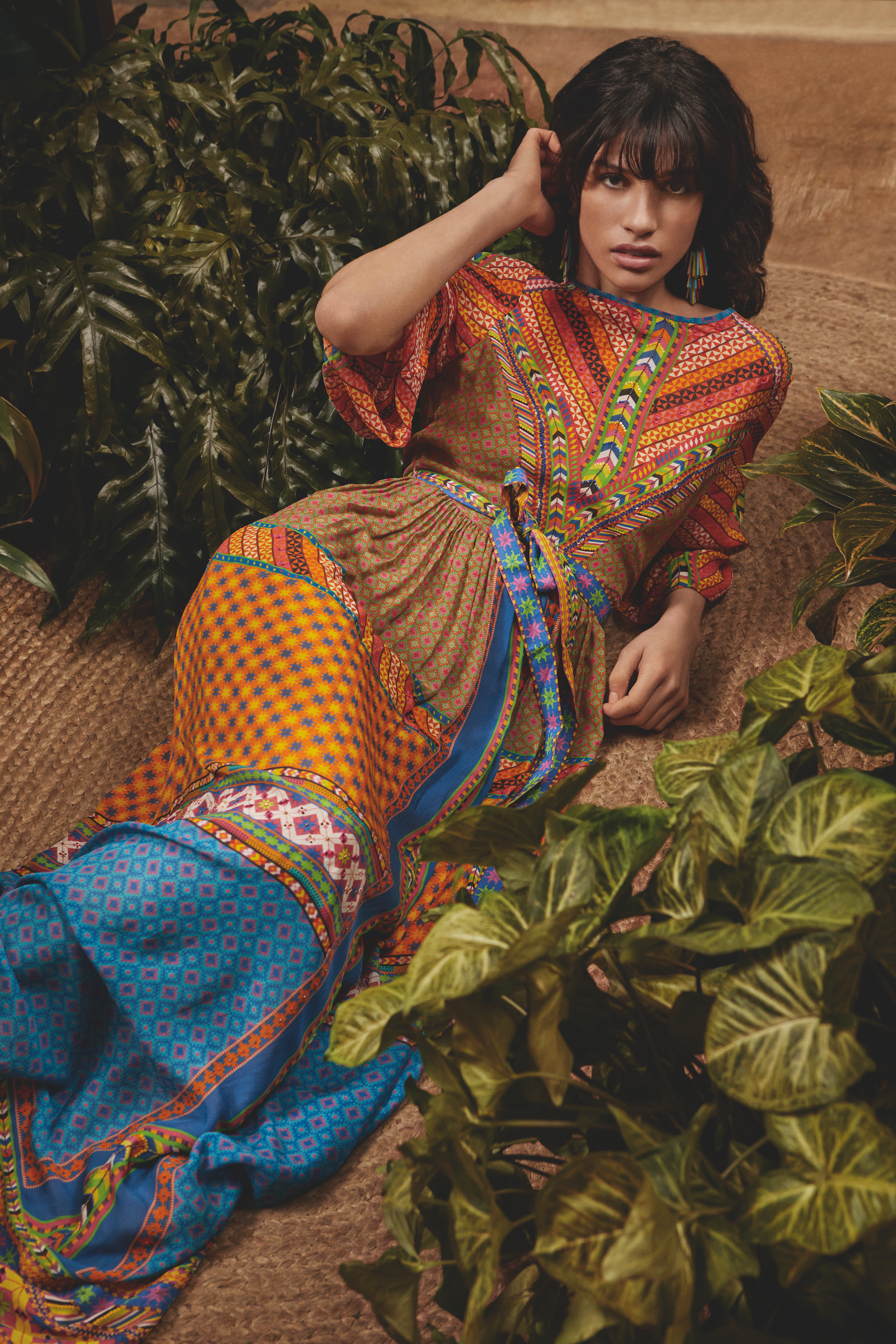A woman with dark wavy hair and bangs lying on a textured beige rug surrounded by green plants, wearing a brightly colored patterned dress with geometric and floral designs.