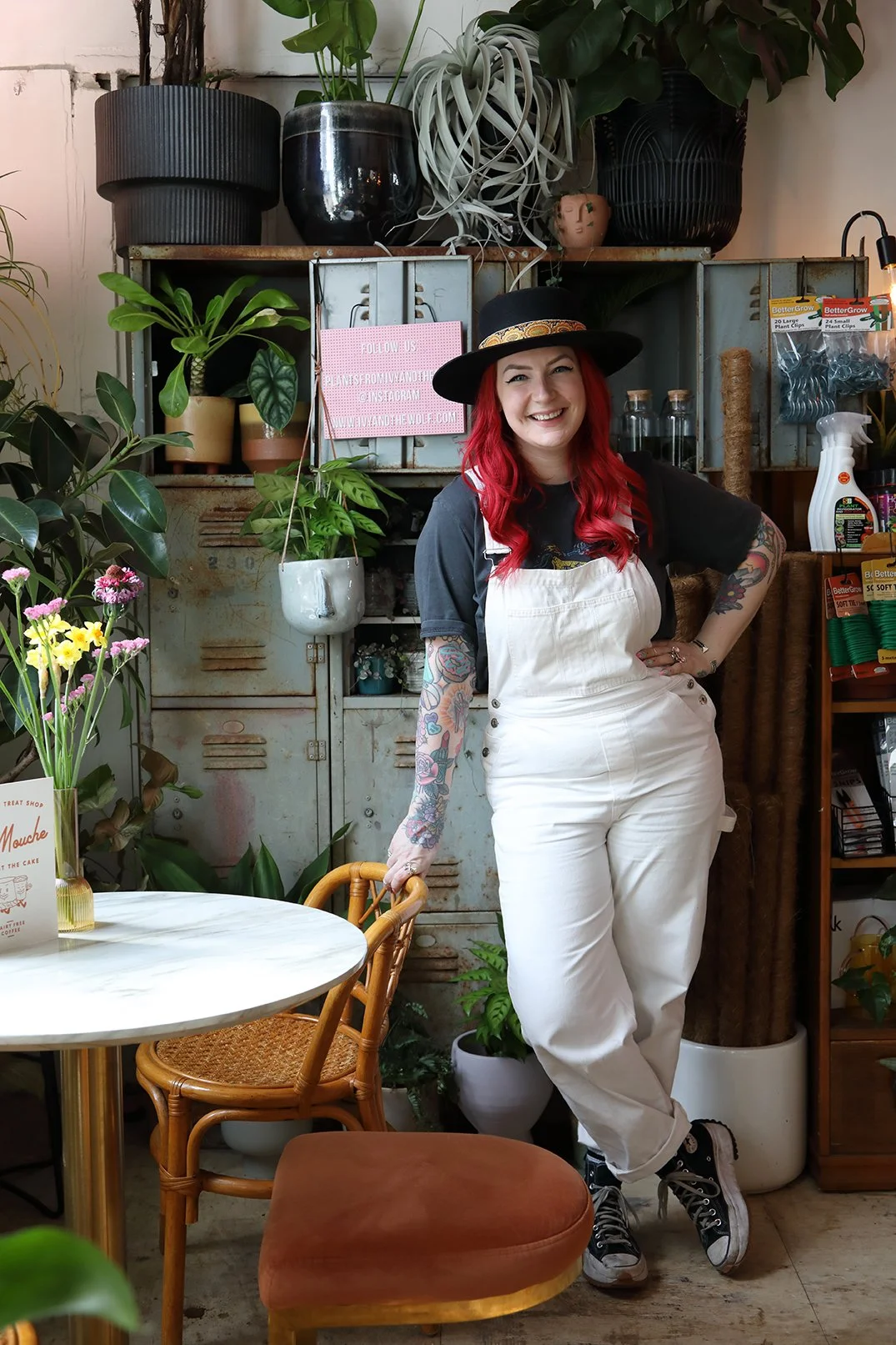 A woman with red hair, tattoos on her arms, wearing a black hat, black shirt, and white overalls, smiling and standing indoors among plants and vintage lockers.