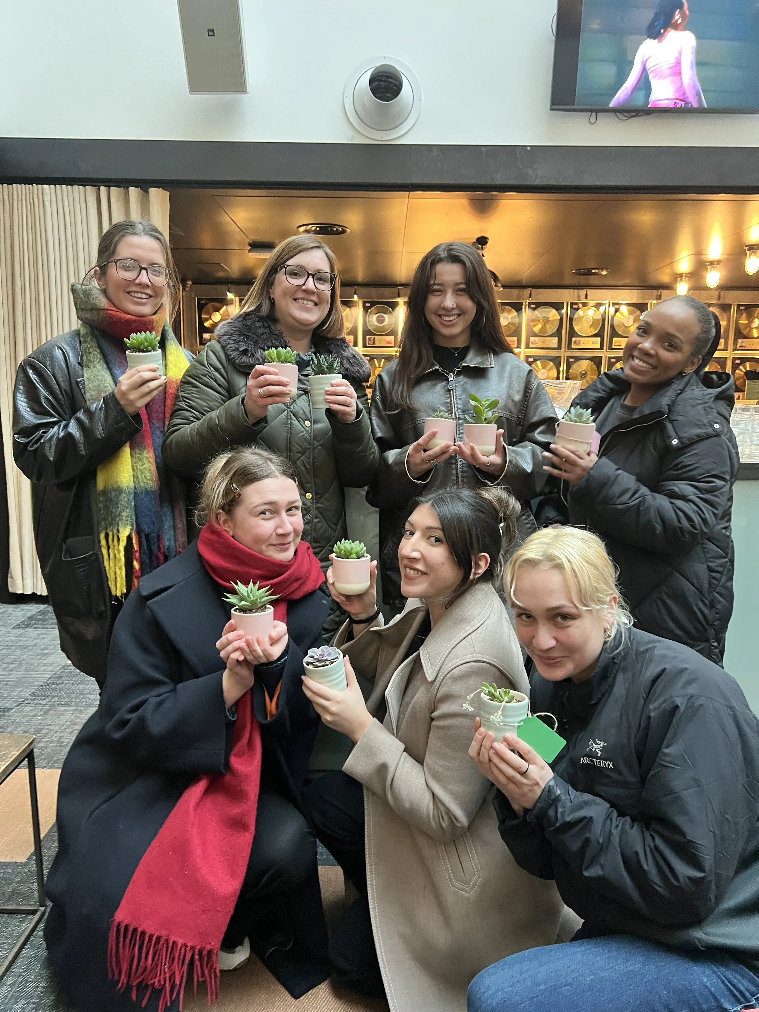 Group of eight women at an indoor location, each holding small potted succulents, smiling for the photo.