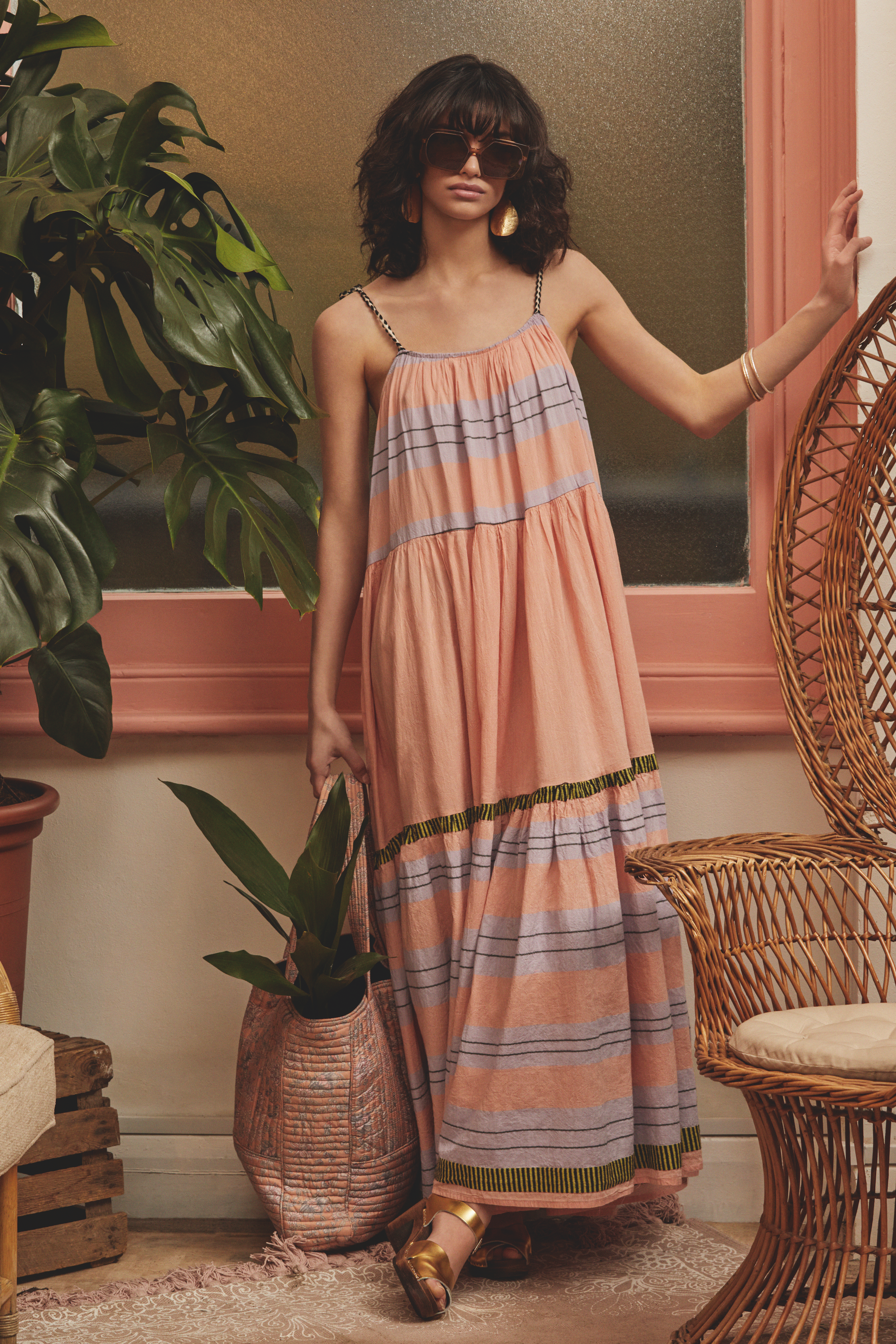 A woman standing indoors wearing sunglasses and a long, colorful, striped summer dress with spaghetti straps. She is holding a potted plant and resting her hand on a wall. The room has rattan furniture, a large leafy plant, and a window with pink tri