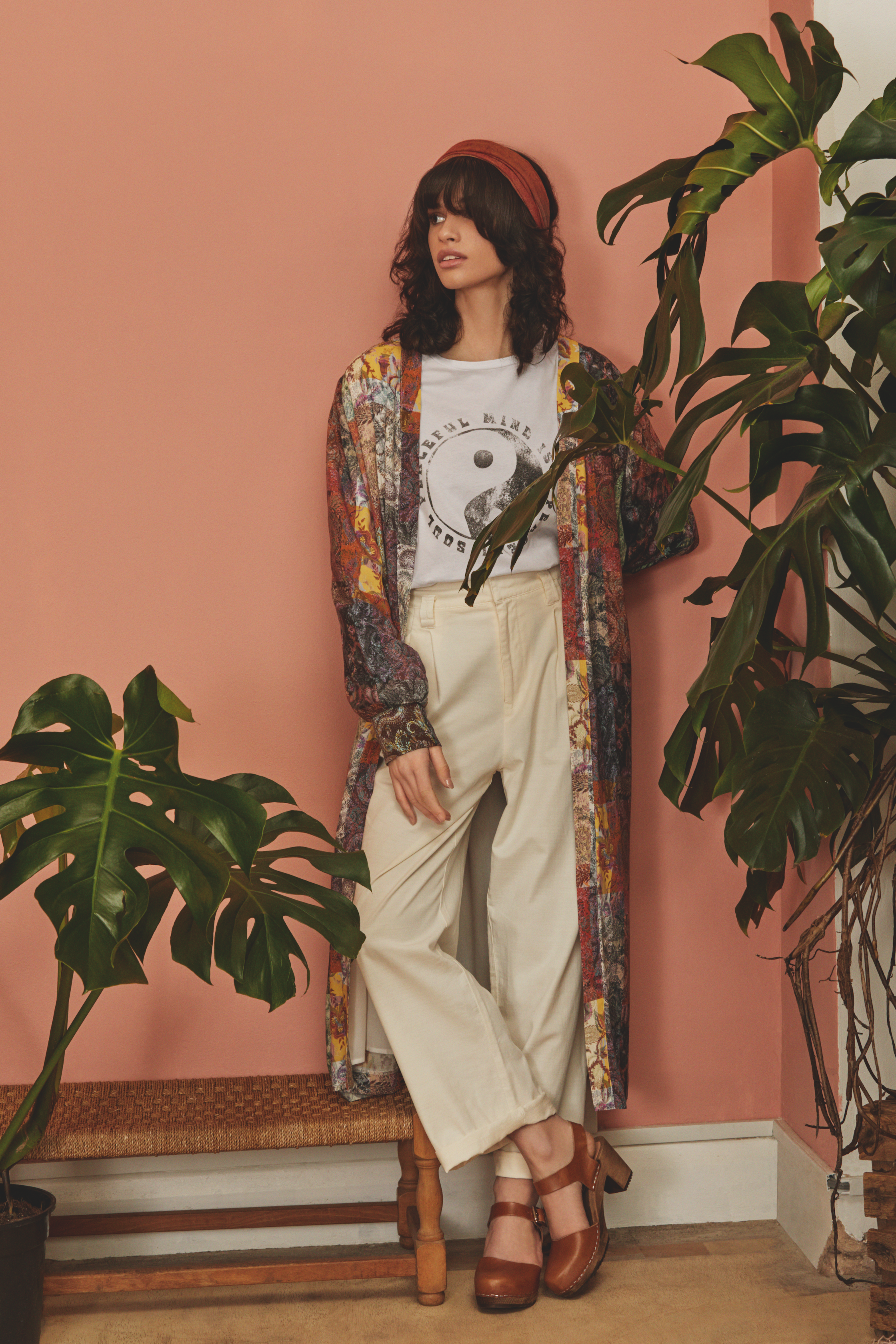 A woman with dark curly hair, wearing a red headband, a colorful patterned long coat, a white graphic t-shirt with Yin-Yang symbol, beige loose pants, and brown platform shoes, standing in a room with pink walls and large green houseplants.