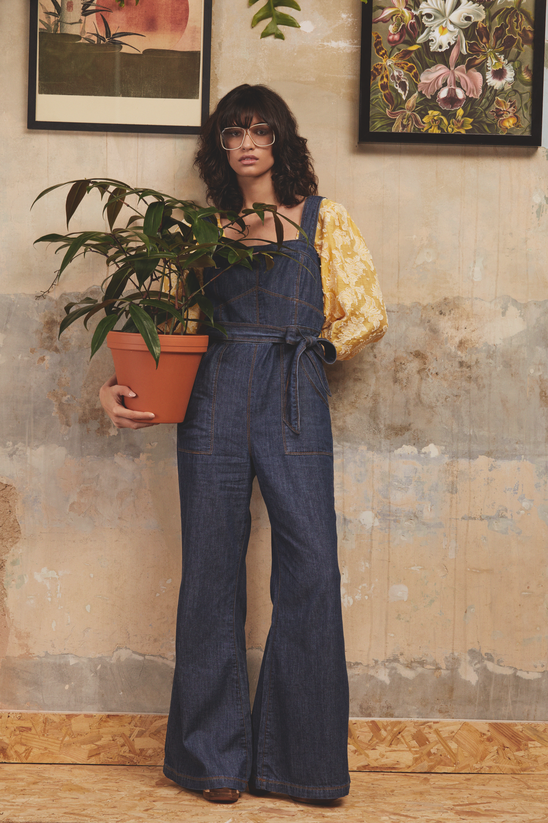 A woman with shoulder-length dark curly hair and glasses stands against a textured beige wall, holding a potted plant with large dark green leaves. Two framed botanical prints hang on the wall behind her, and she wears a yellow floral blouse under de