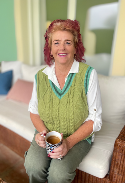A woman with curly red hair sitting on a white cushioned bench, holding a mug of tea, in a room with green walls and colorful cushions.