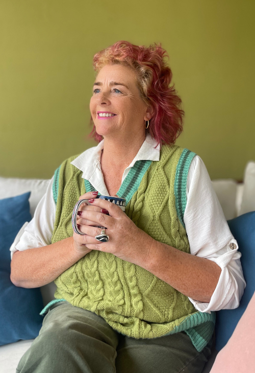 A smiling woman with short red hair and earrings, sitting on a couch holding a mug, wearing a white shirt and a green vest, with a green wall background.