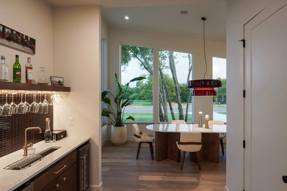 Interior view of a modern dining area with large windows showing trees outside. There is a round dining table with six chairs, a large potted plant, and a contemporary red ceiling light fixture. Adjacent to the dining area, a small bar or refreshment station has a white countertop, glassware, bottles, and a sink.