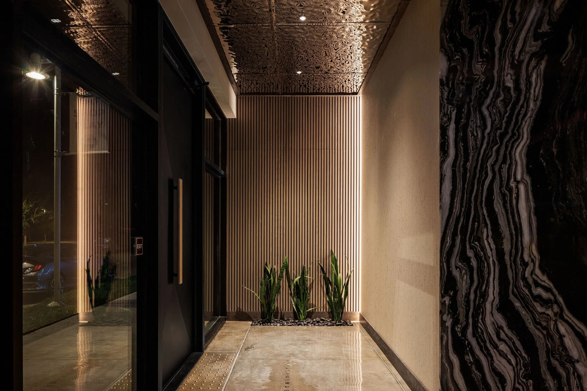Modern interior hallway with black sliding door, wooden wall paneling, and decorative marble wall. Green plants in a small bed against the wooden wall. Recessed ceiling lights and polished concrete floor.