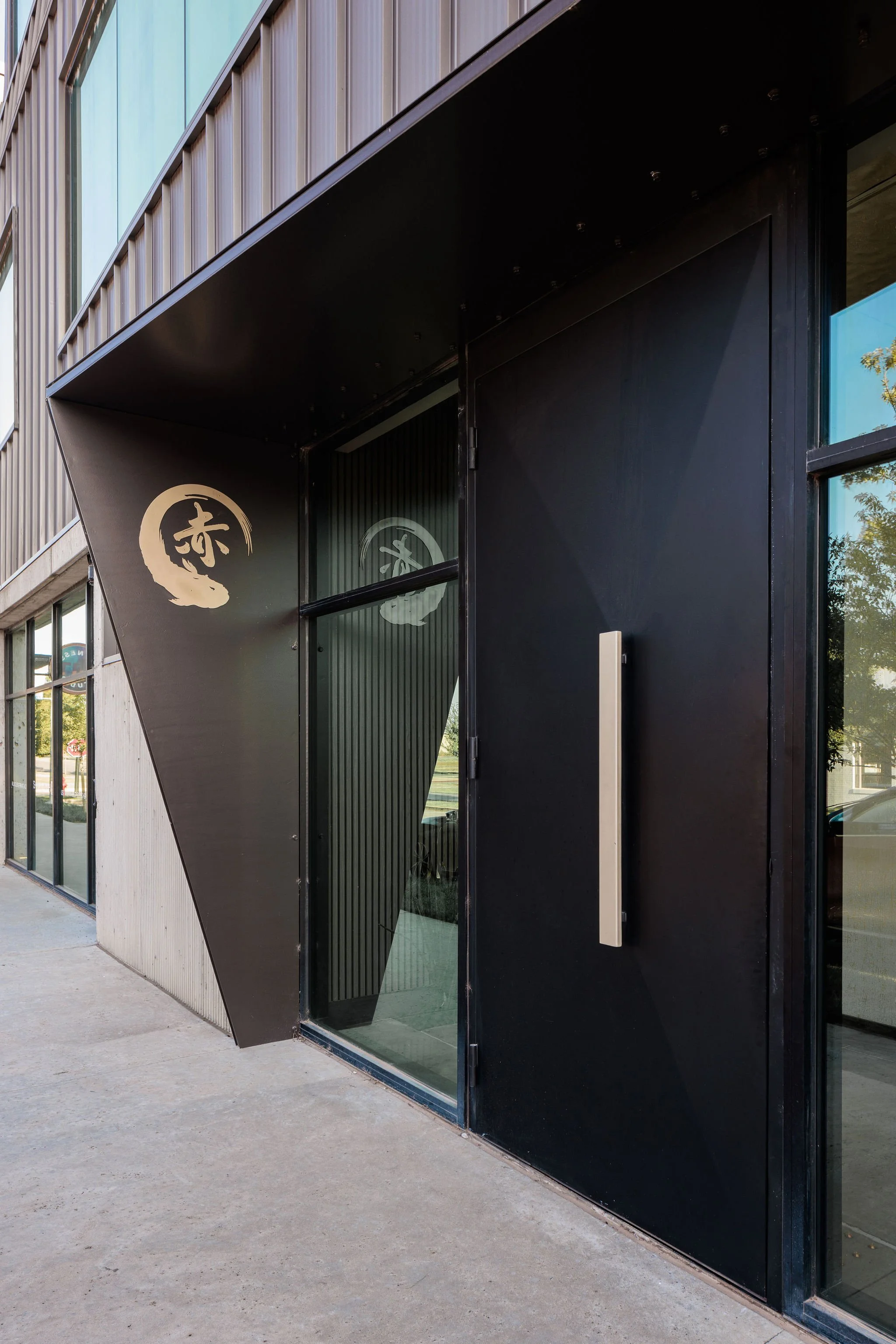 Modern storefront with black exterior, large metal handle, glass door, and Japanese-style logo featuring a white brushstroke circle with Japanese characters inside.