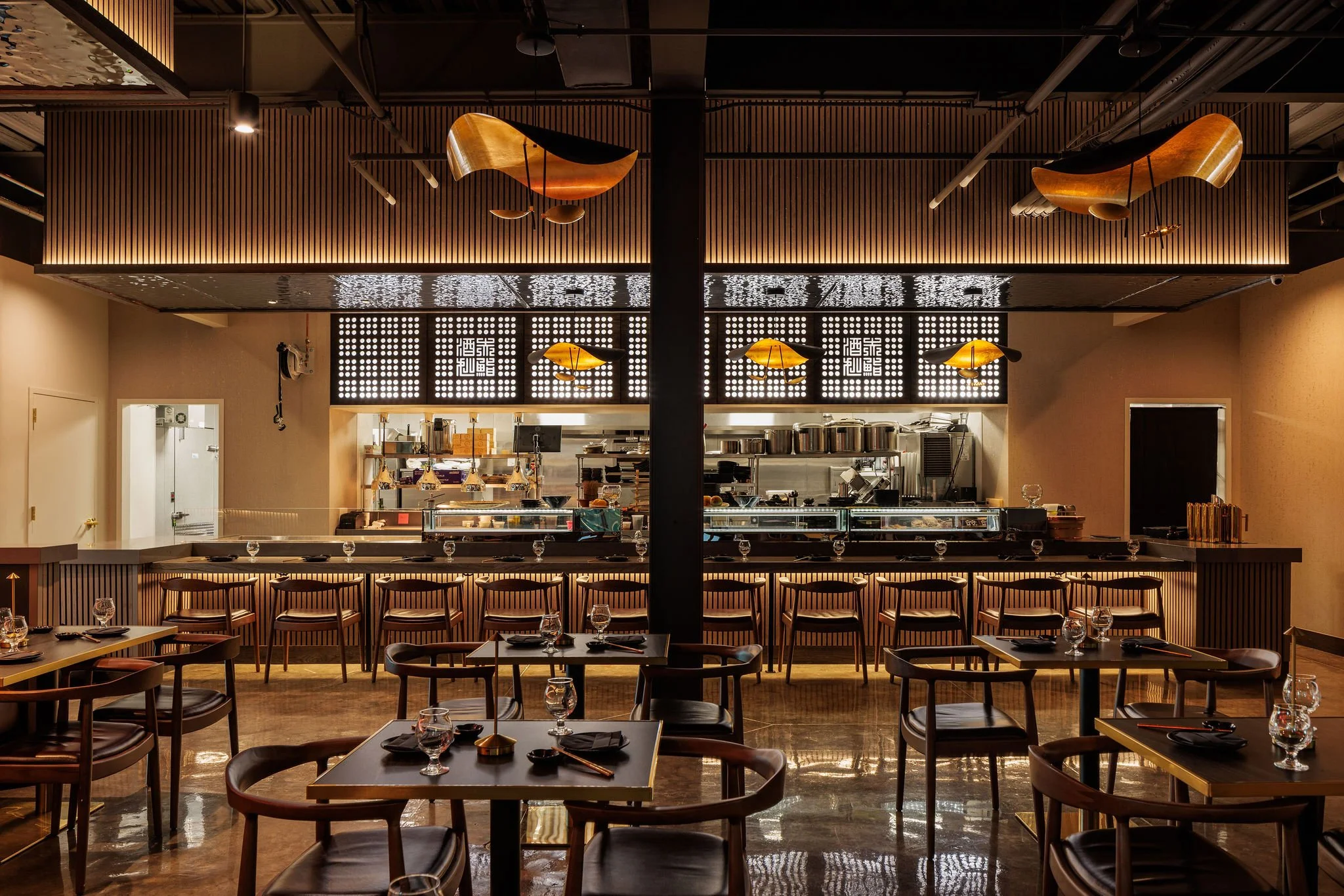 Interior of a modern restaurant or cafe with a bar counter, wooden chairs, and tables set with glassware and black napkins. Decorative wooden ceiling fixtures and pendant lights hang above, and the open kitchen area is visible in the background.