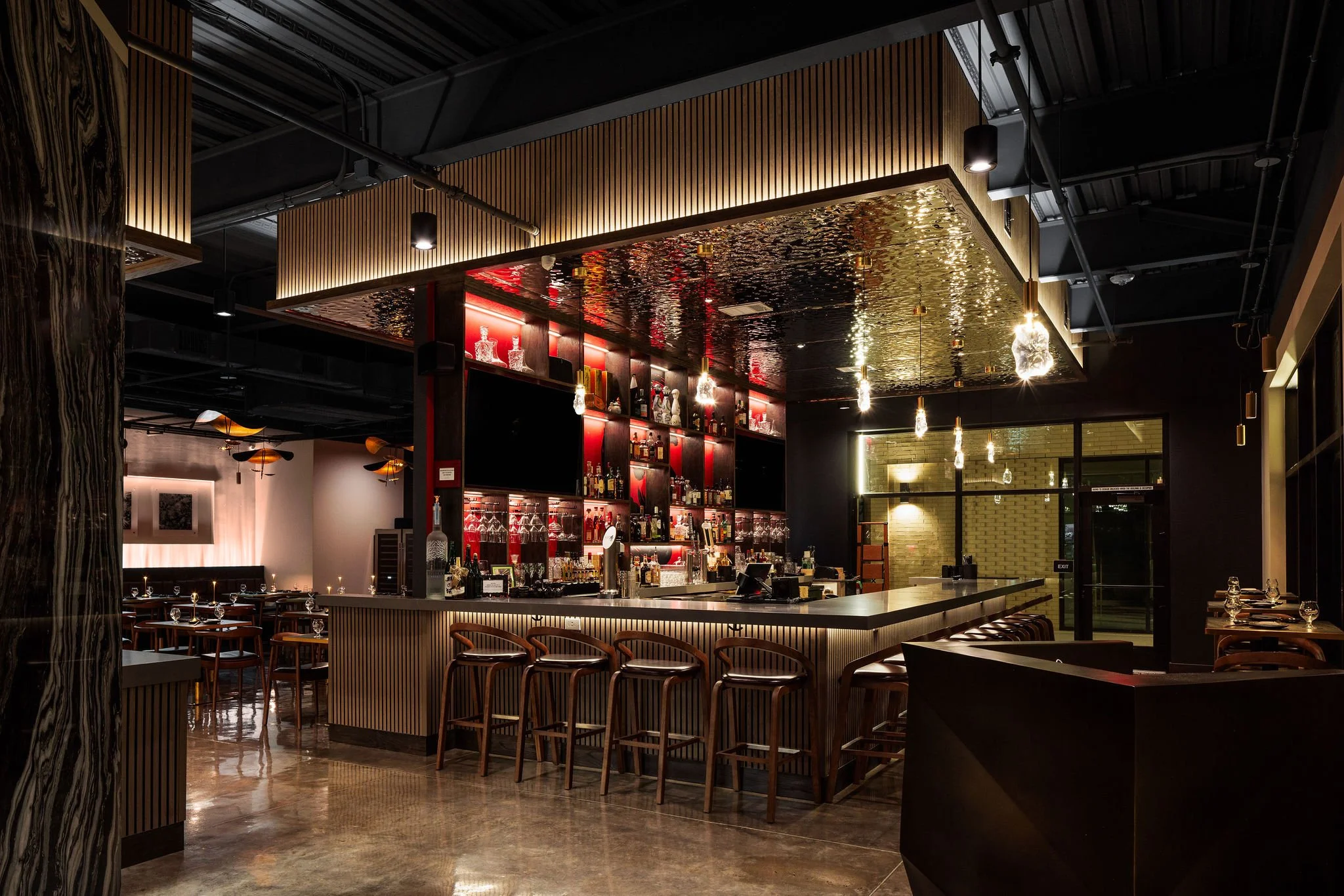 Modern bar with dark walls, wooden accents, and warm lighting, featuring a curved bar counter and shelves stocked with bottles.