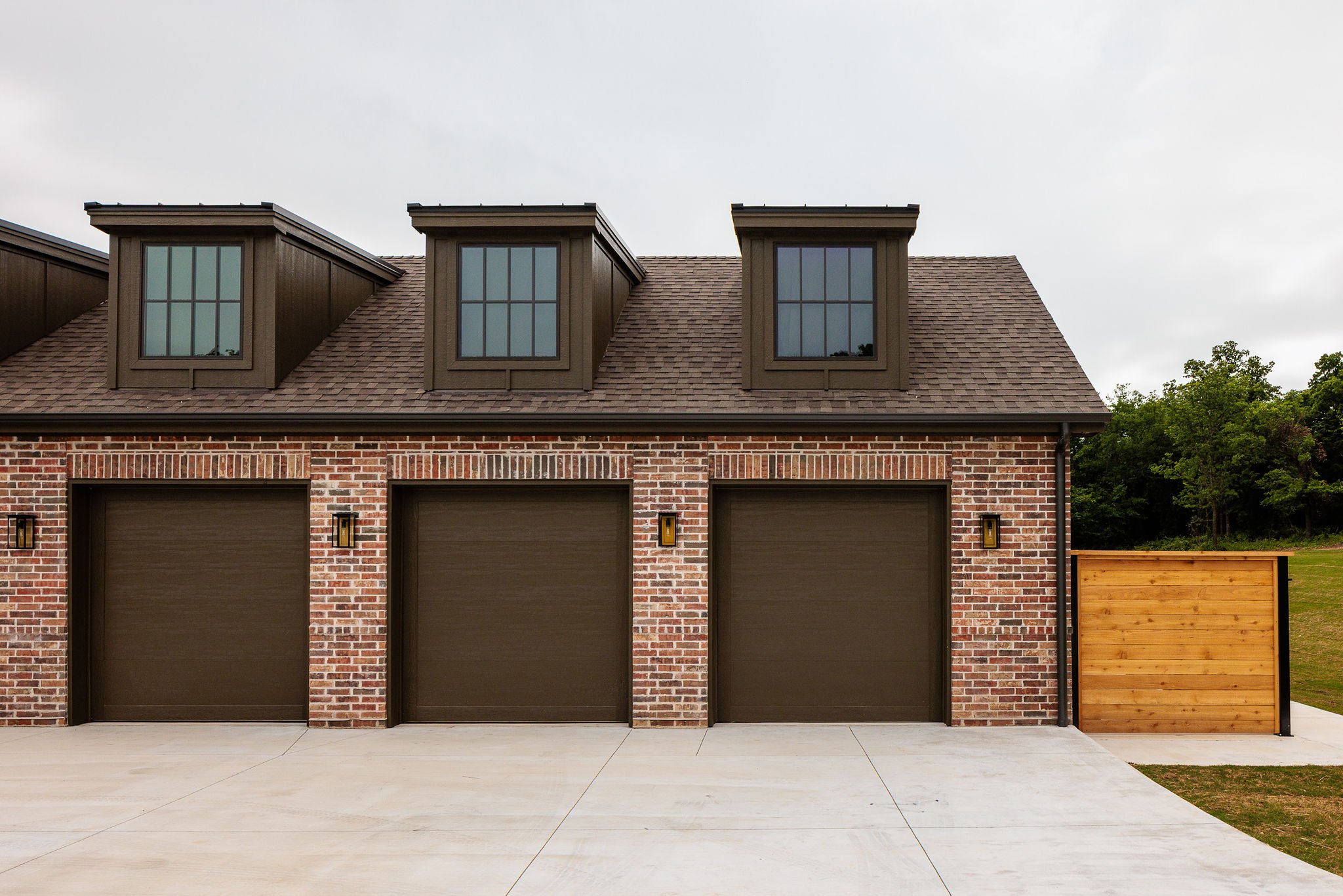 Front view of a house with three garage doors, brick exterior, three dormer windows on the roof, and a small wooden fence on the right side.