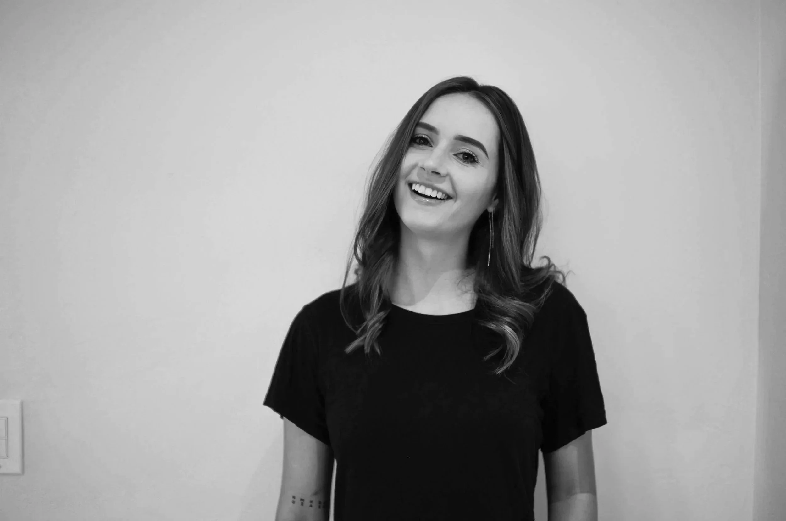 A smiling woman with wavy hair wearing a black t-shirt standing against a plain wall.