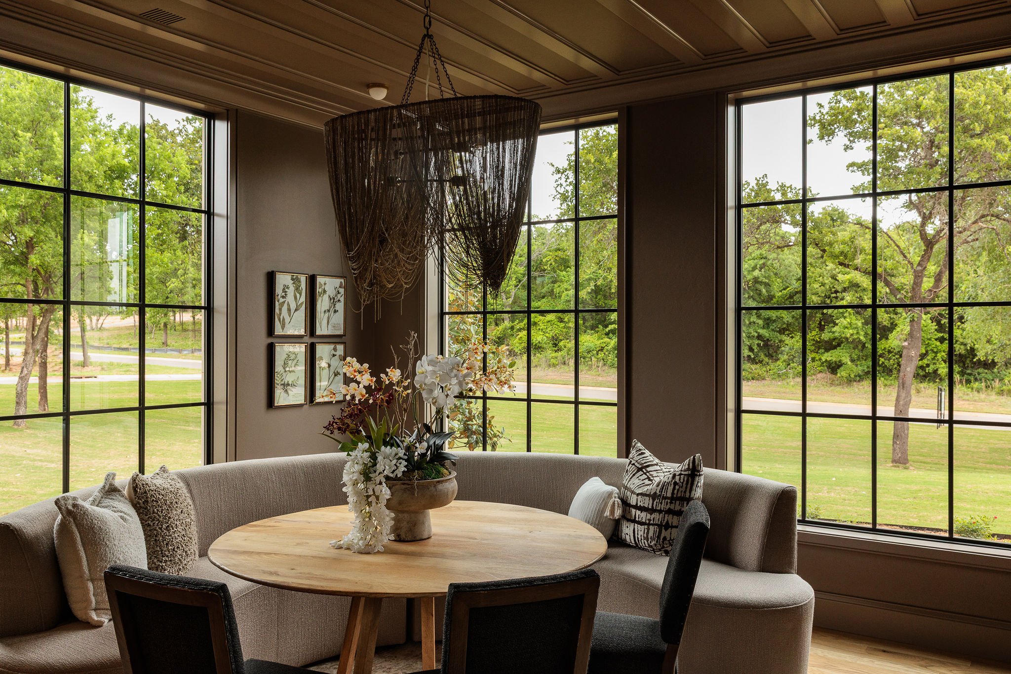 A cozy living room with large windows showing a green outdoor landscape, a round wooden table with a floral centerpiece, and a curved sofa with cushions.