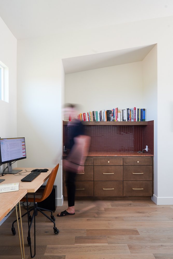 Blurred person with dark hair wearing black pants, pink top, and black sandals, standing in a room with a wooden desk, Apple monitor, and a bookshelf on a brown cabinet with six drawers. The room has a light-colored wooden floor and white walls, with a small window visible to the left.