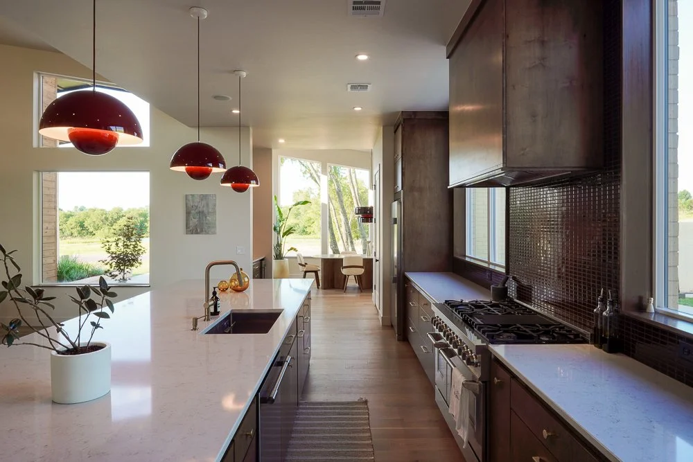 Modern kitchen with large island, dark wood cabinets, stainless steel stove, and pendant lights. Large windows show greenery outside.