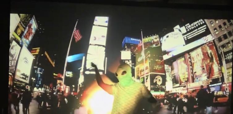 An alien figure wearing a green face mask waving in Times Square at night with illuminated billboards and people walking on the street.