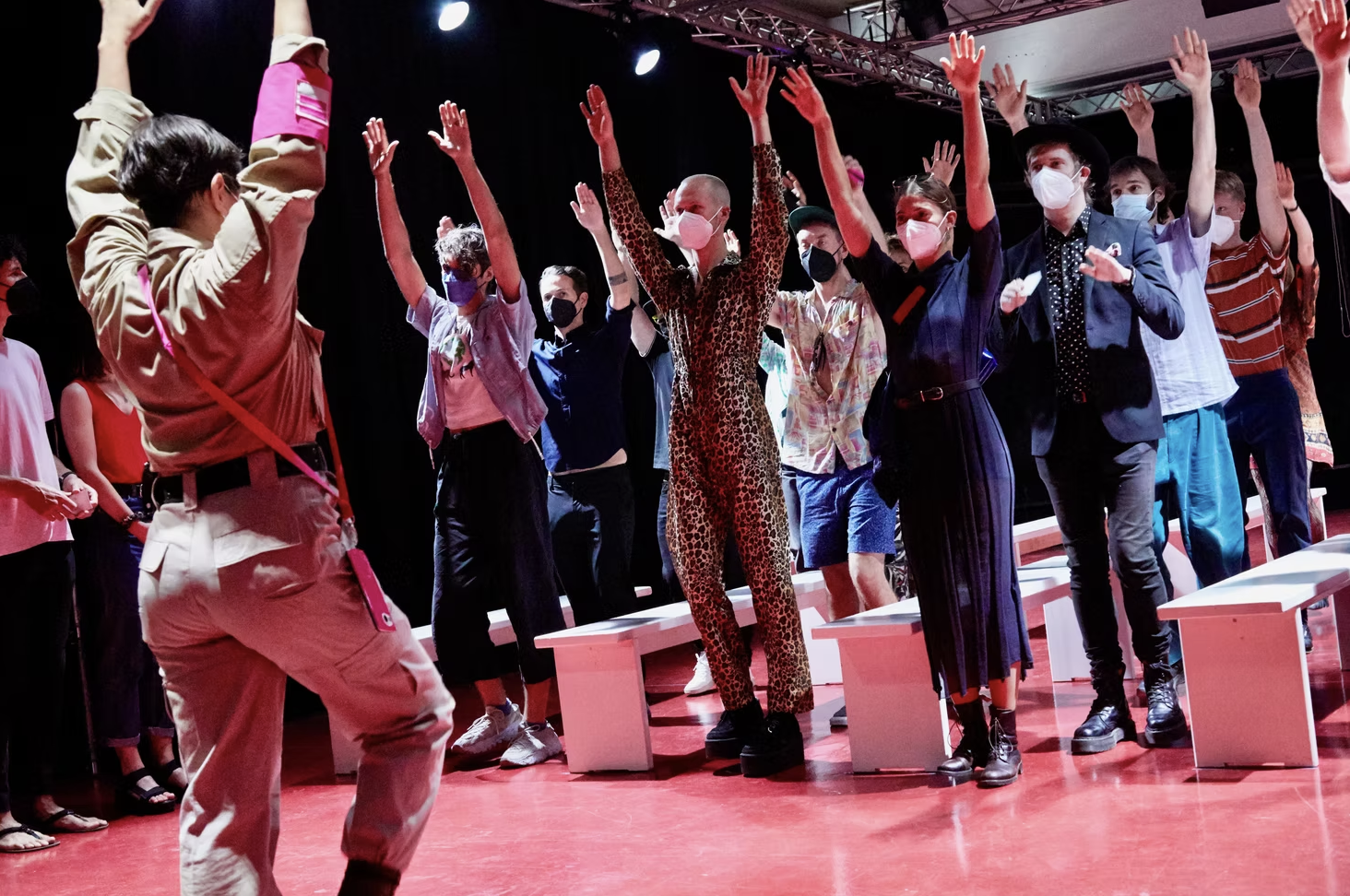 People standing and raising their hands, participating in an activity or performance, with some wearing masks, in a room with red flooring and benches.