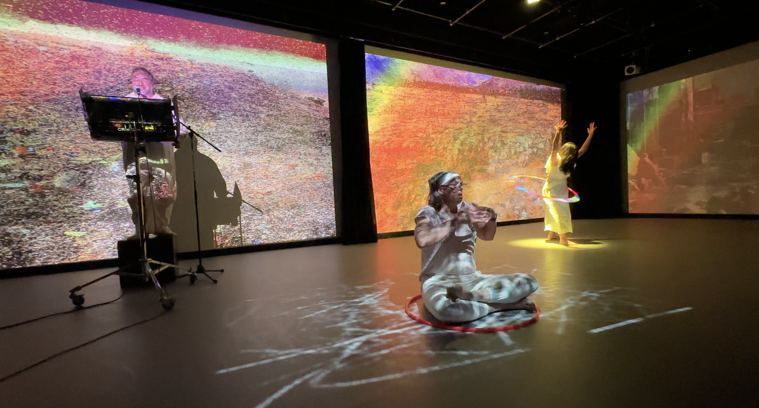 Three performers on a dark stage with digital backdrops displaying colorful, abstract, and digital-like images. One person is sitting cross-legged on the floor with a hula hoop around them. Another stands with arms raised, also with a hula hoop, and the third stands at a microphone, possibly singing or speaking.