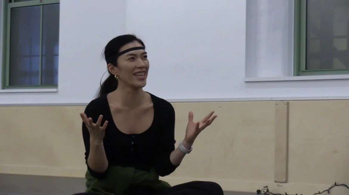 A woman with long dark hair, wearing a black top, gold earrings, and a headband, smiling and gesturing with her hands in a casual indoor setting near windows.