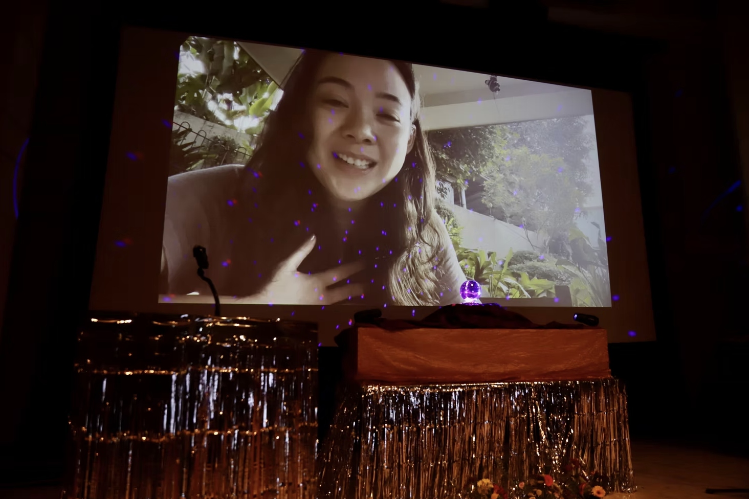 A woman smiling during a video call projected on a screen, decorated with a sparkling tablecloth and small decorative items, with purple laser lights and a small purple crystal ball on a table.