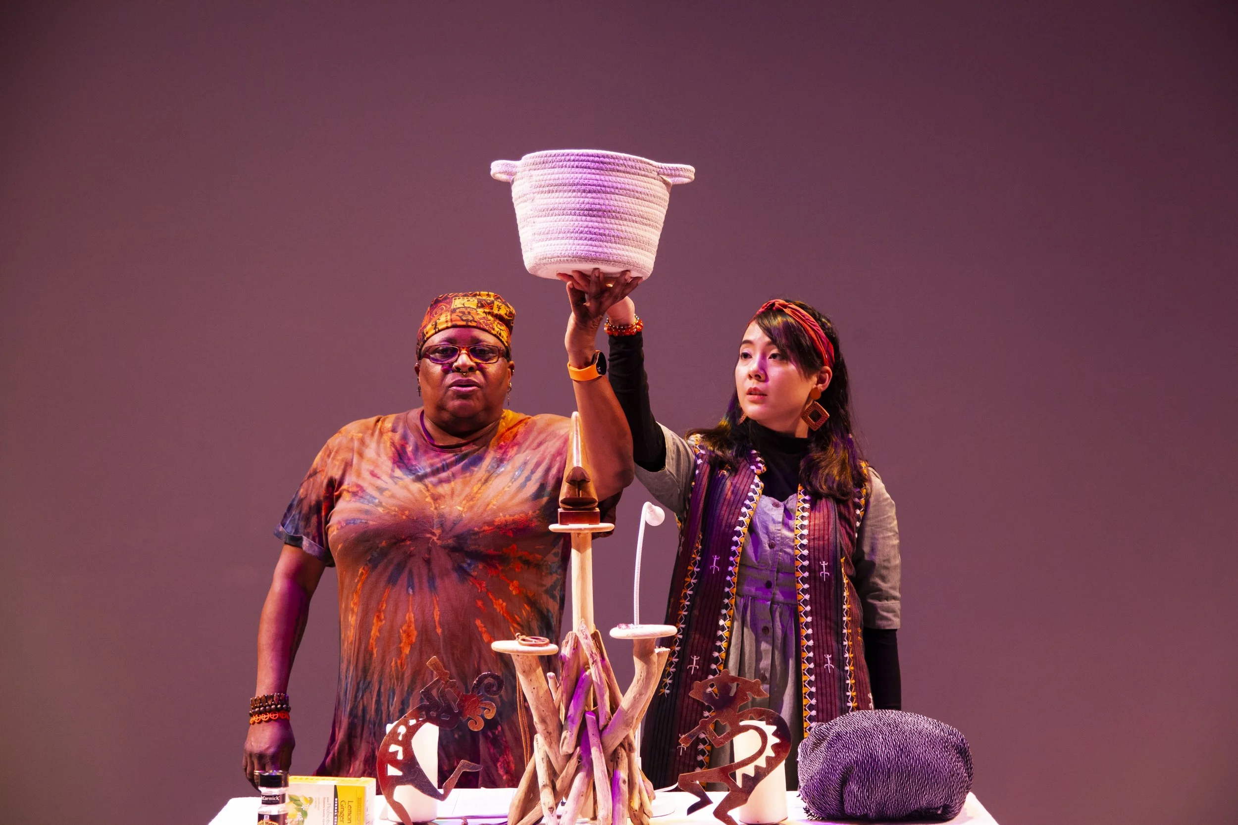 Two women stand behind a table holding a woven basket high. The woman on the left is wearing glasses, a multicolored tie-dye shirt, and a headscarf. The woman on the right has long dark hair, is wearing a patterned vest, earrings, and wristbands. The table has various decorative items and containers.