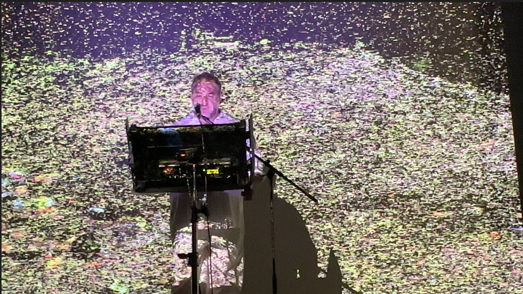 A person performing with a microphone on stage, with a colorful, abstract background projection.